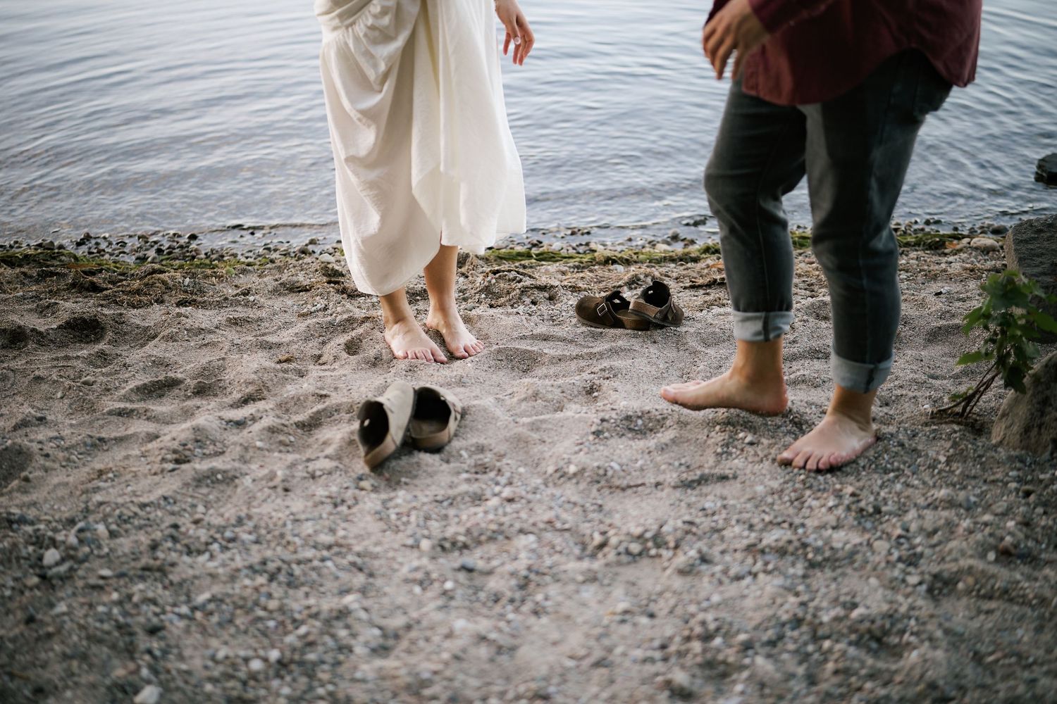 Bare feet and shoes on a sandy beach shore with calm water in the background.