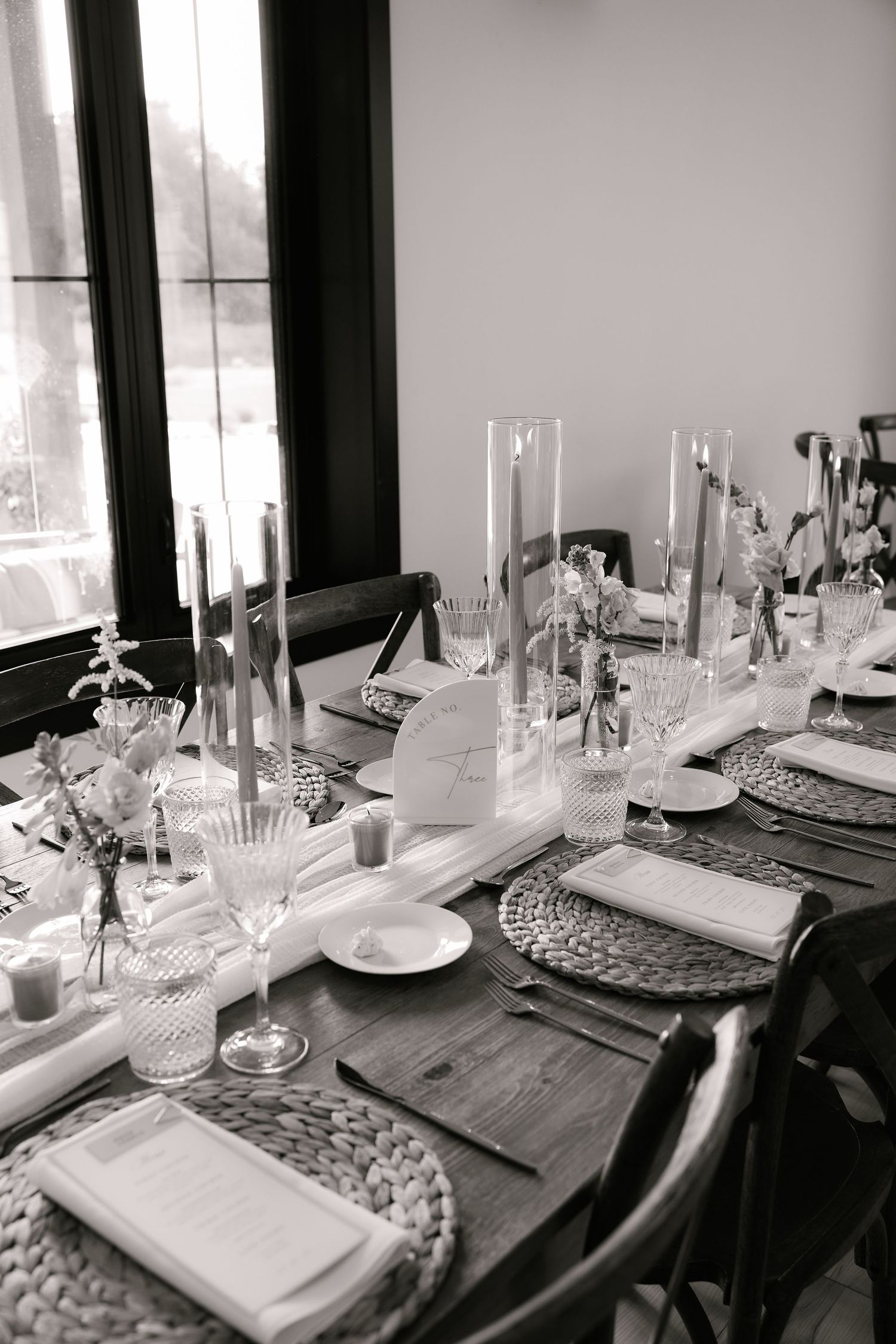 Black and white photo of a rustic wooden banquet table elegantly set with candles, crystal glasses, and woven placemats.