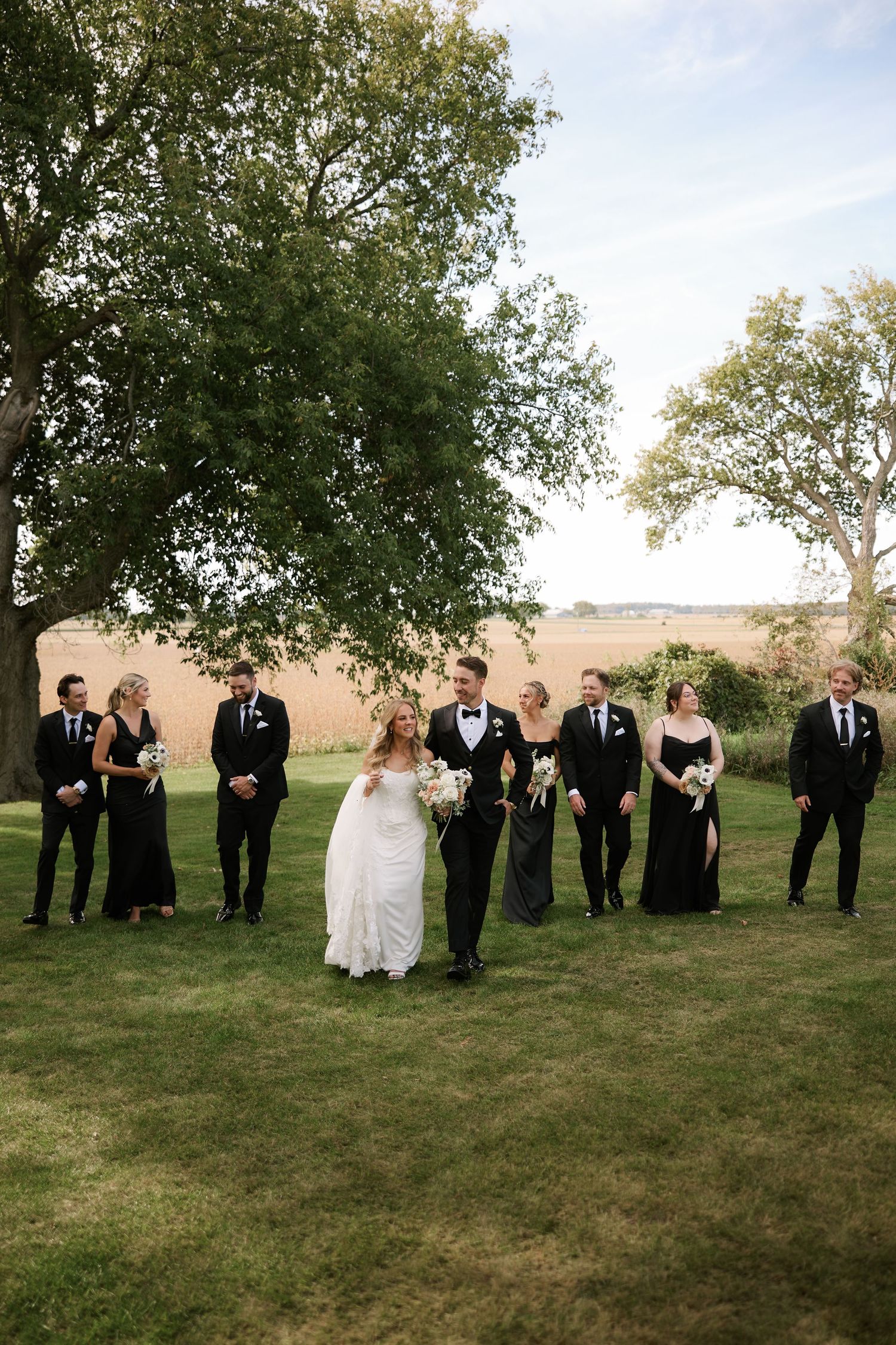 Wedding party in black attire walks with bride and groom on green lawn with trees and golden fields backdrop.