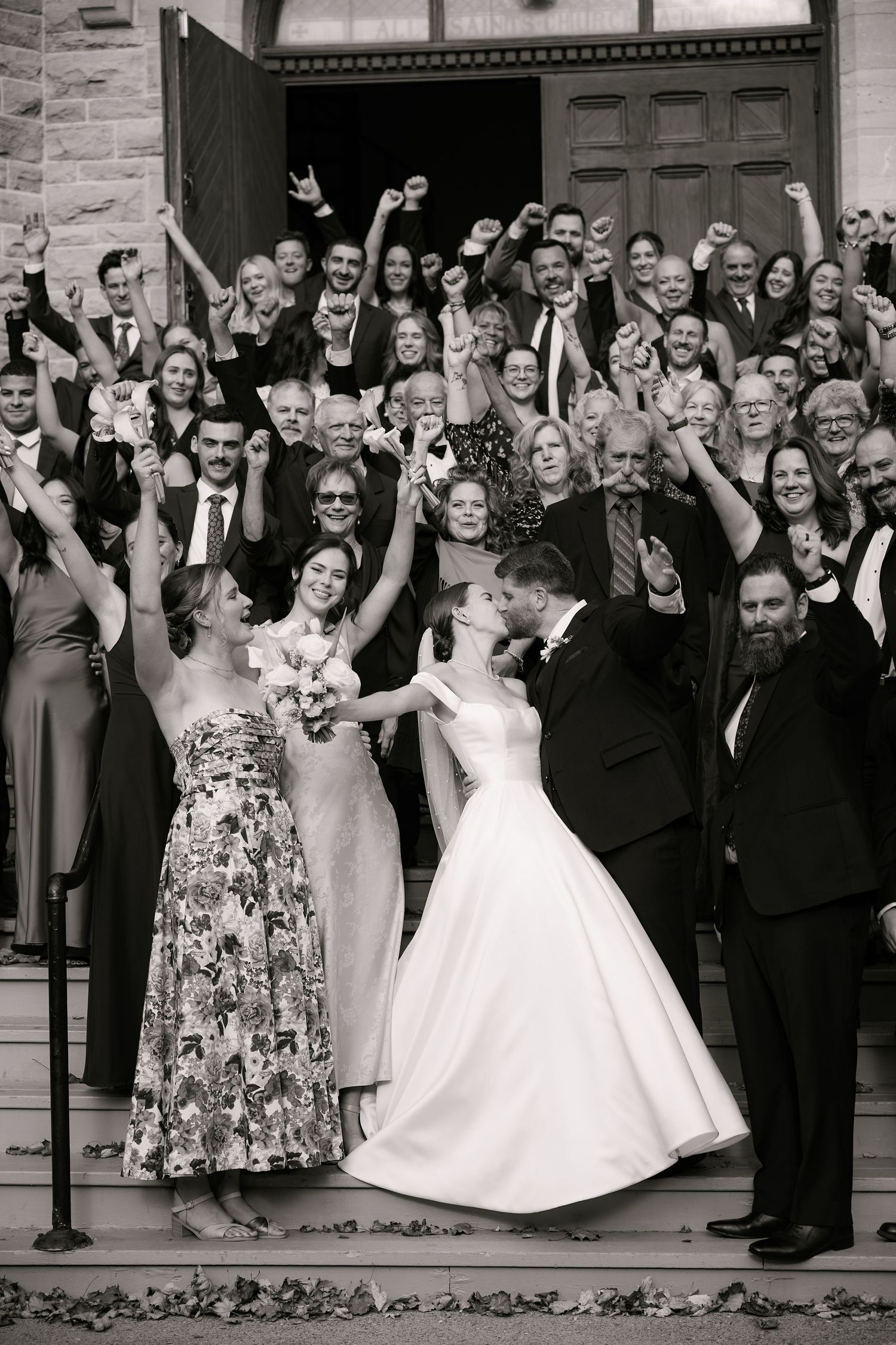 Black and white photo of bride and groom kissing on church steps surrounded by cheering wedding guests.