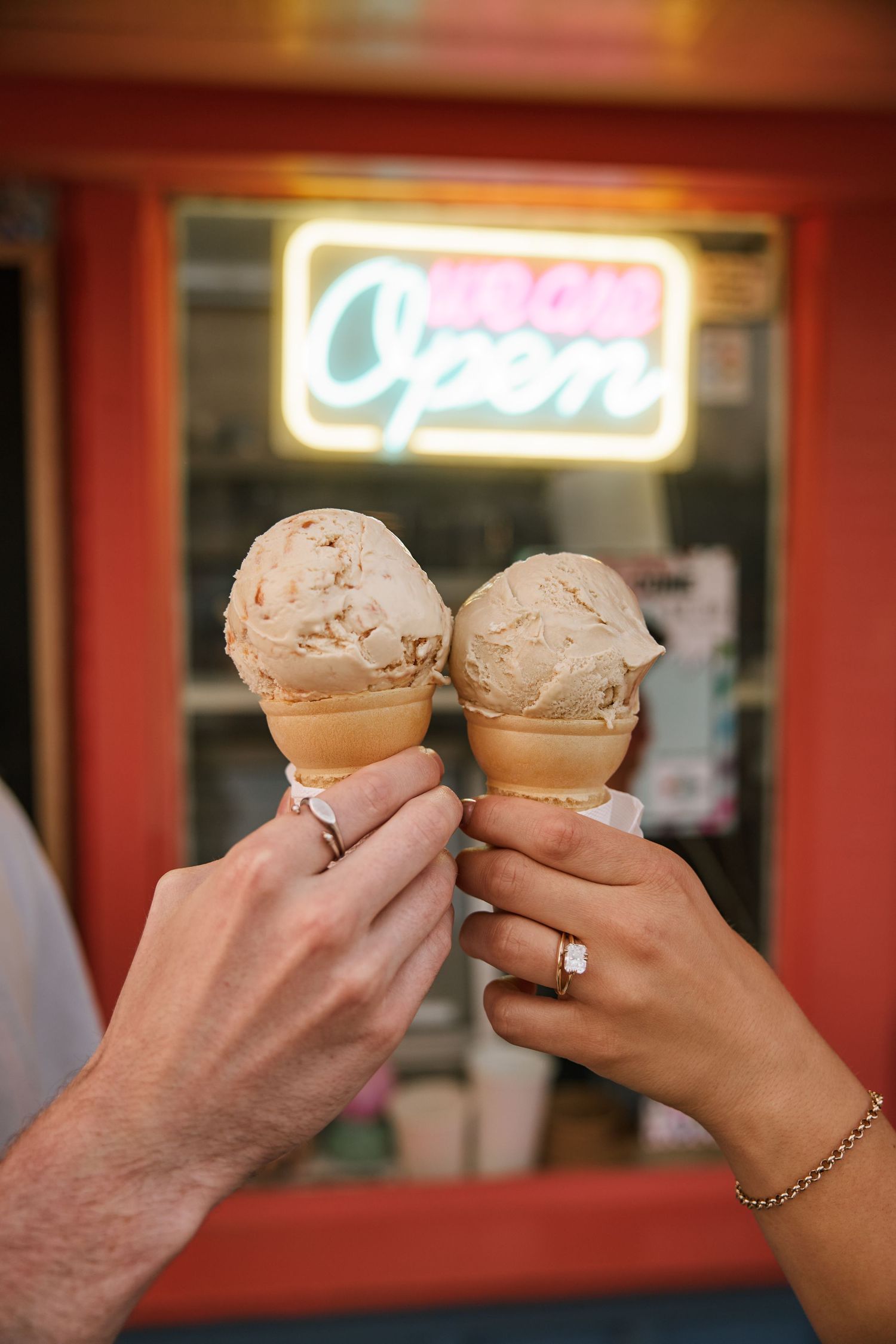 Two hands holding ice cream cones in front of a neon Open sign at an ice cream shop.
