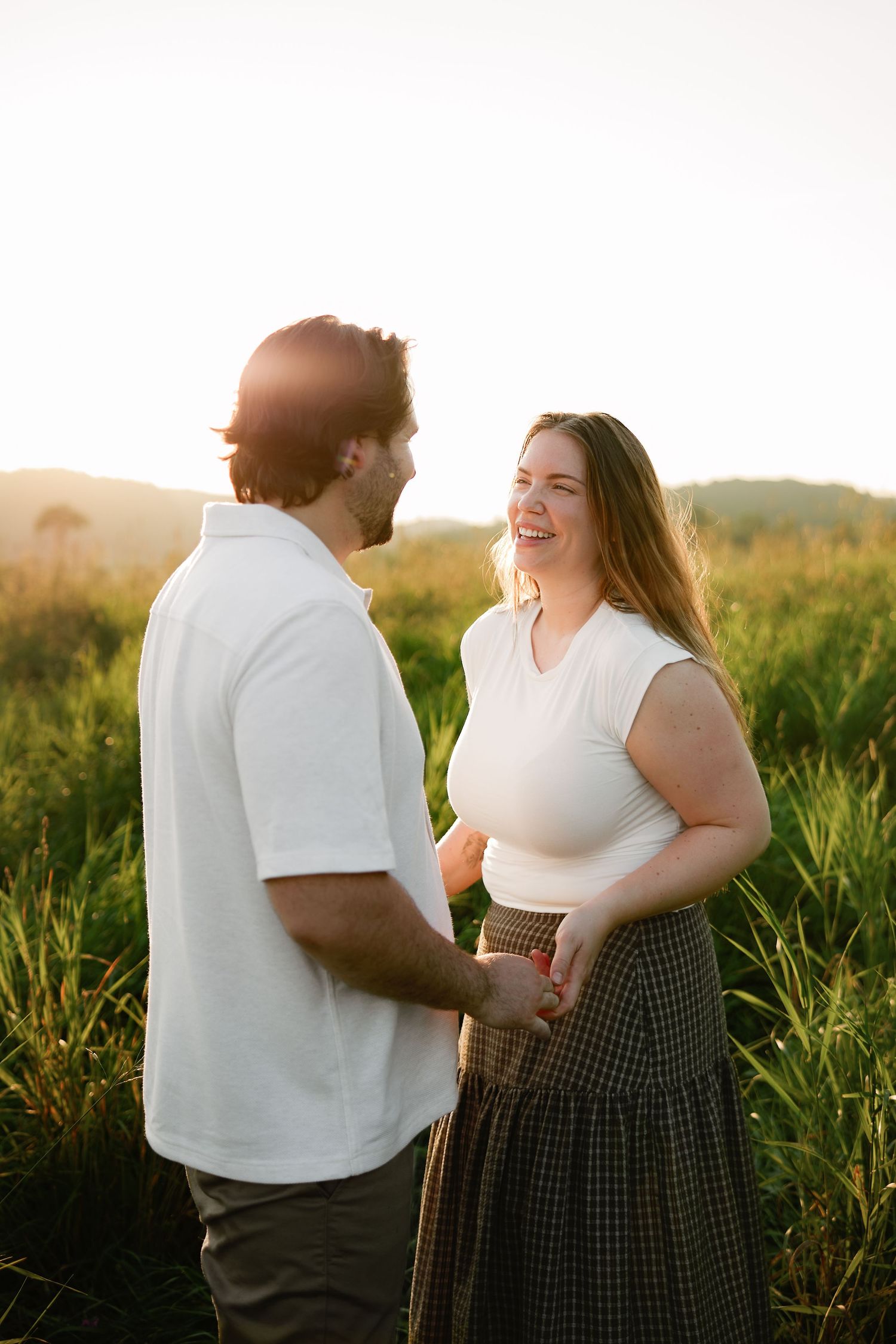 Couple holding hands and laughing together in a golden sunset field, wearing white tops and casual attire.