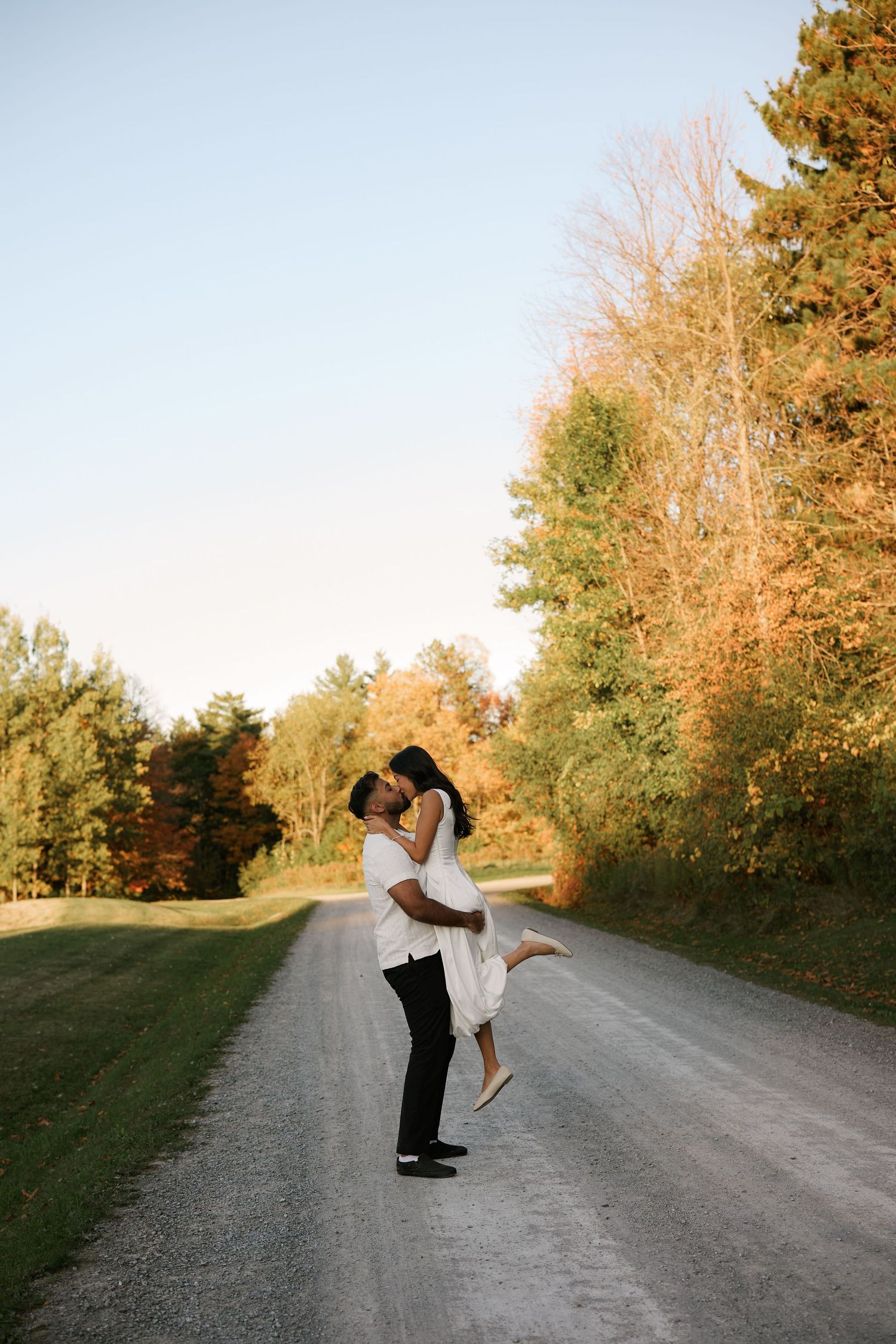 Couple sharing a romantic kiss on a scenic gravel road surrounded by autumn foliage at golden hour.