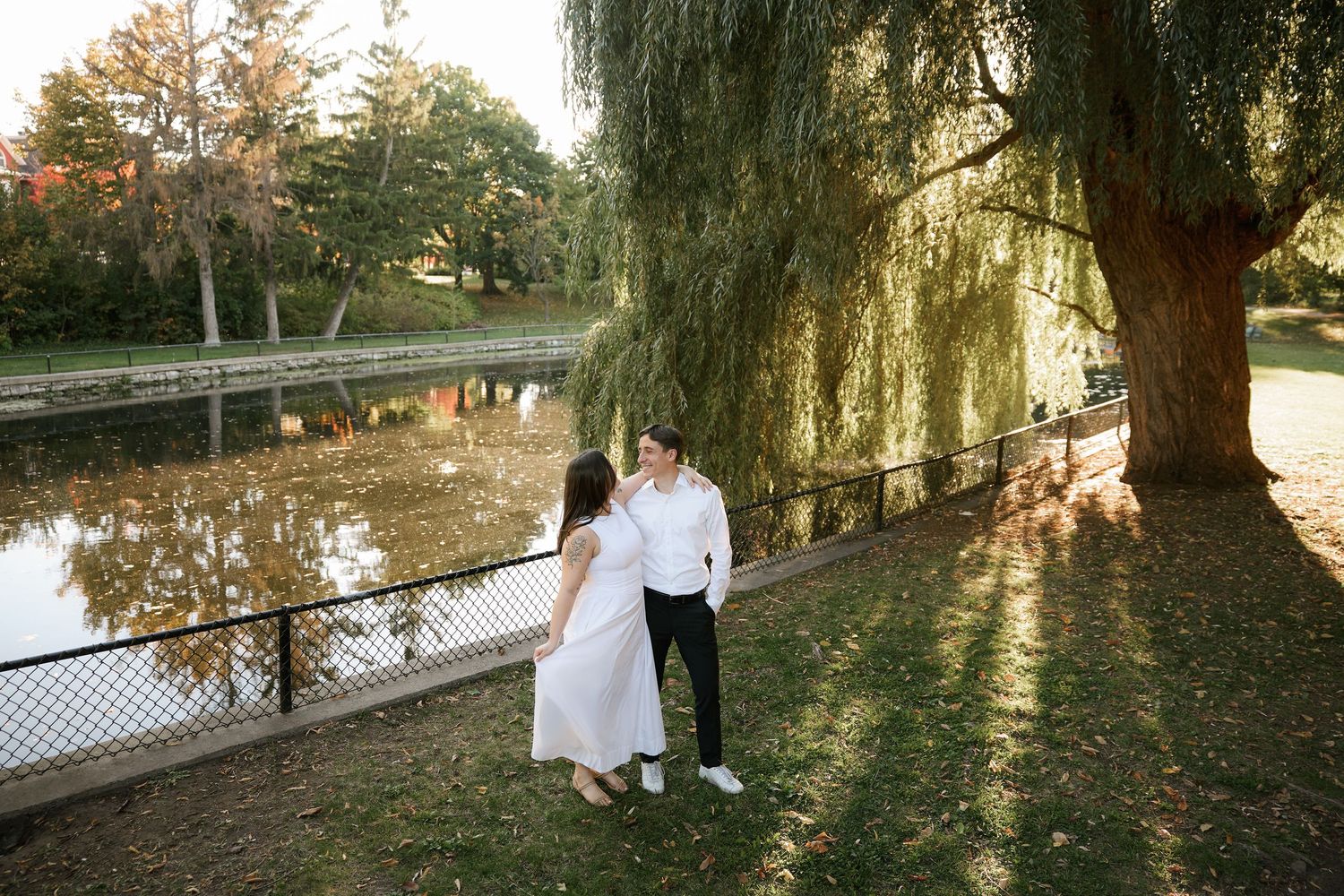Couple holding hands by a serene pond under a weeping willow tree in a lush park during golden hour.