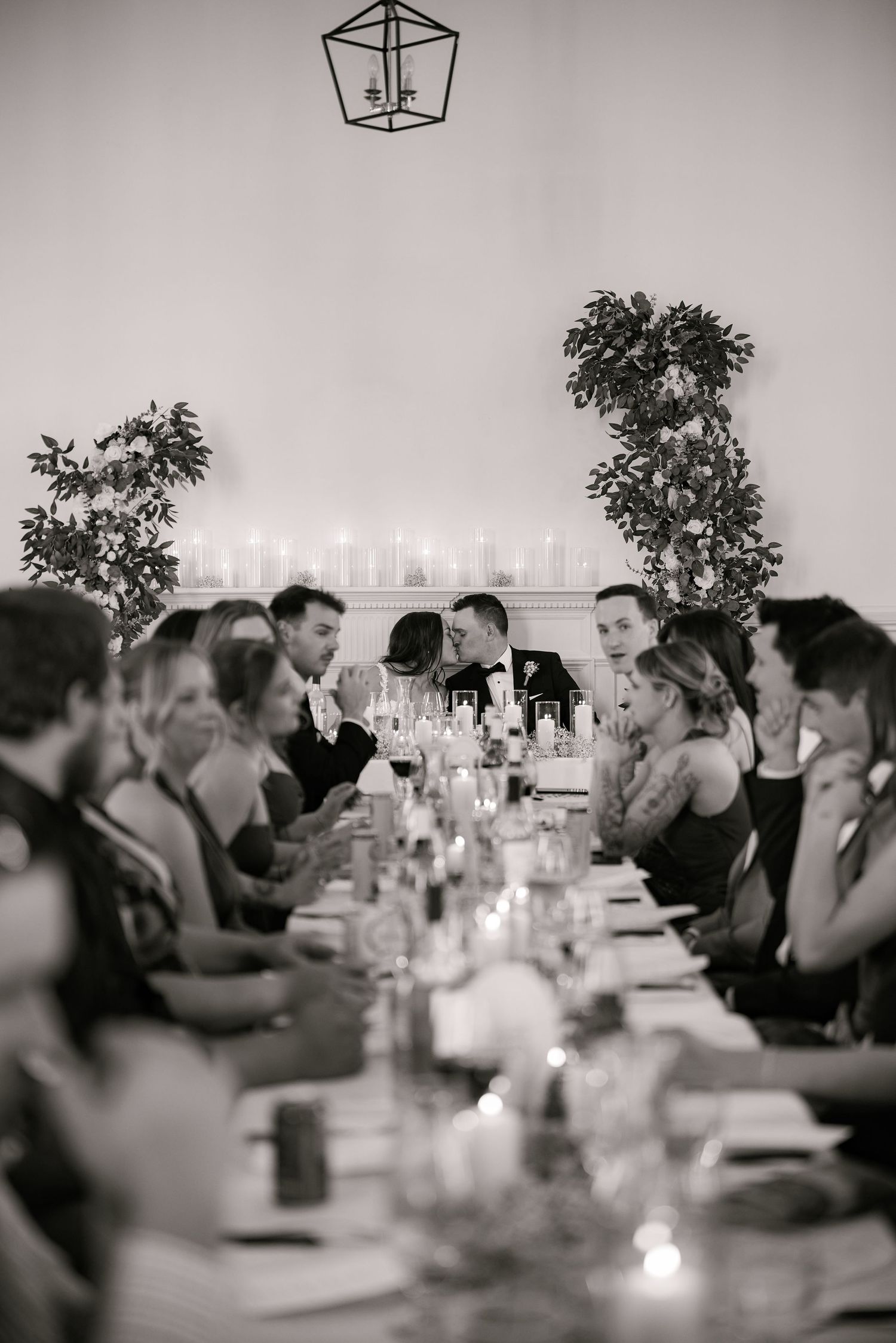 Black and white wedding reception photo of couple kissing at long candlelit table surrounded by guests celebrating.