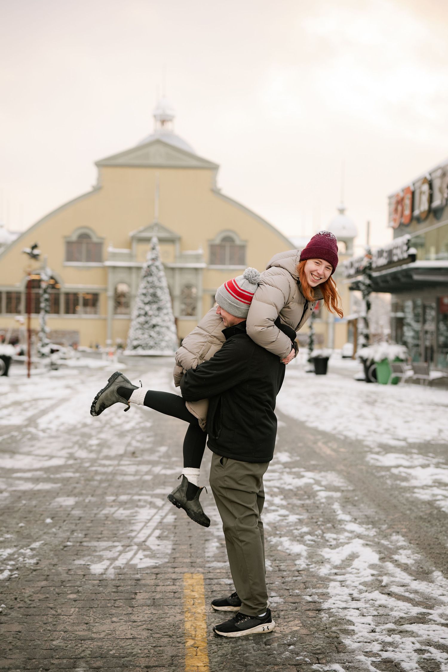 A playful winter scene outside a snow-covered building with a couple embracing in warm winter clothing.