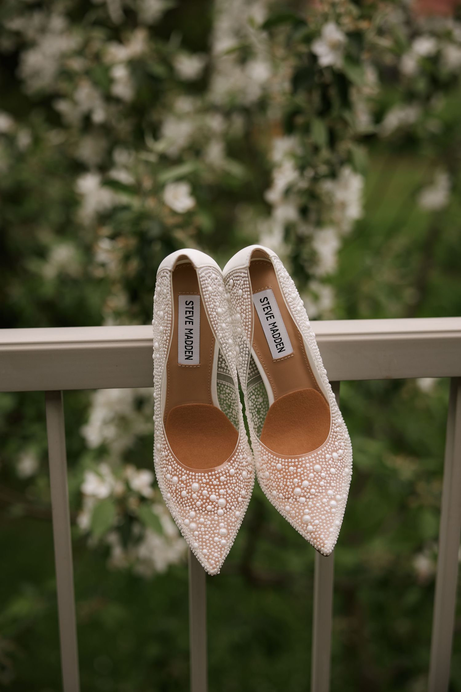 Sparkly embellished wedding shoes displayed on a white railing with blossoming trees in background.
