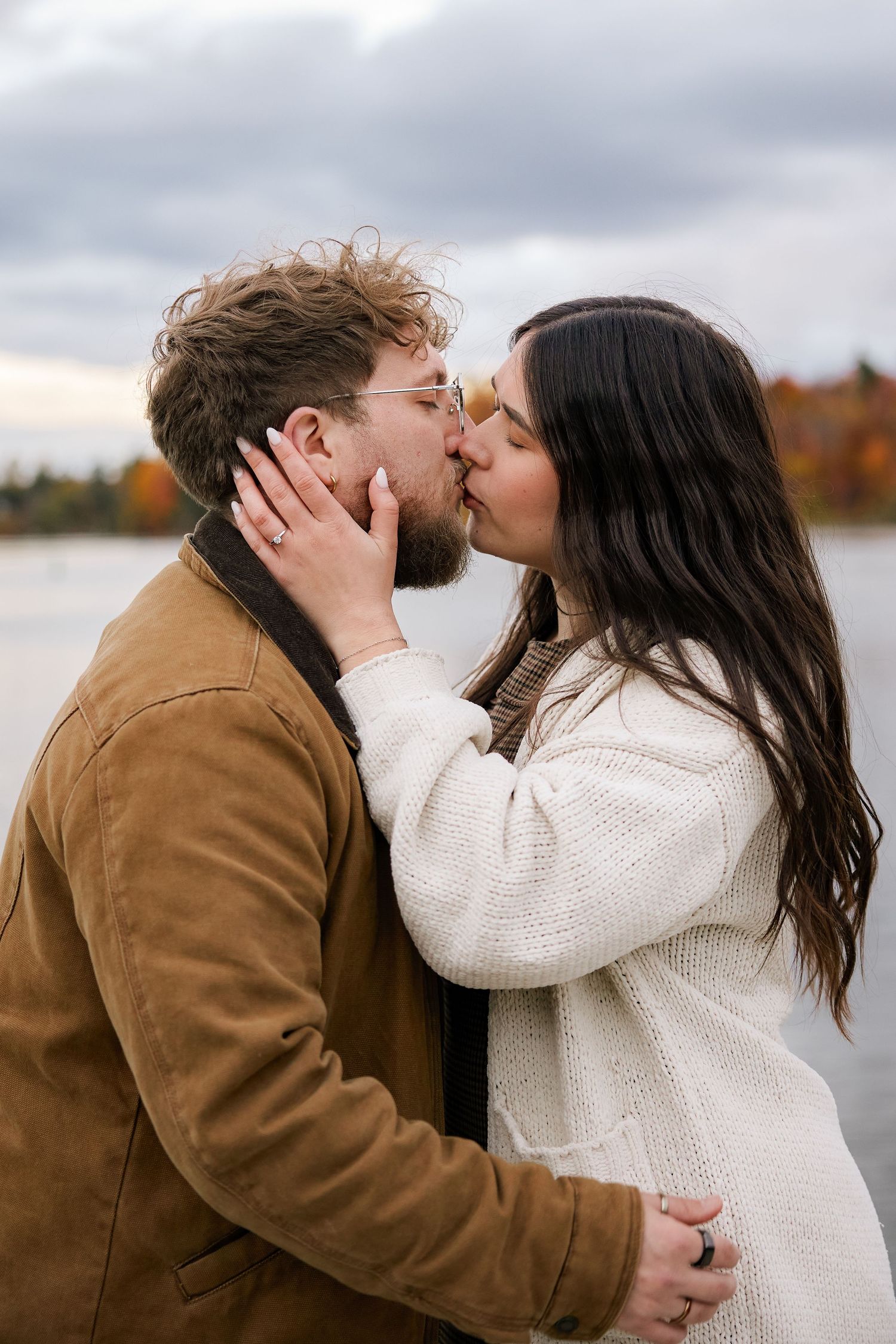 Couple sharing a romantic kiss outdoors by a lake in autumn, wearing cozy sweater and brown jacket.