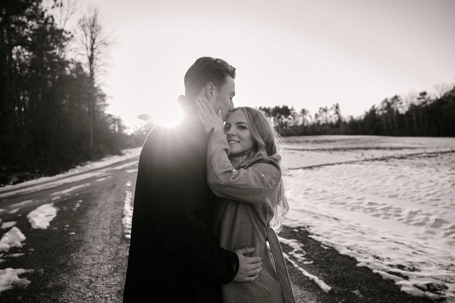 Black and white photo of a couple embracing on a snowy road at sunset with trees in the background.