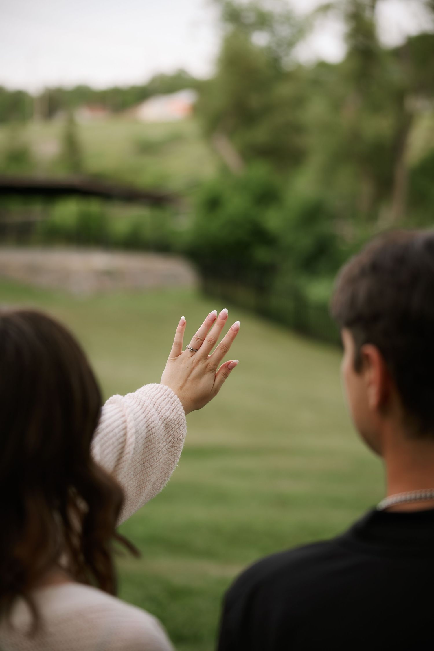 Couple standing outdoors showing off engagement ring on raised hand with green landscape in background.