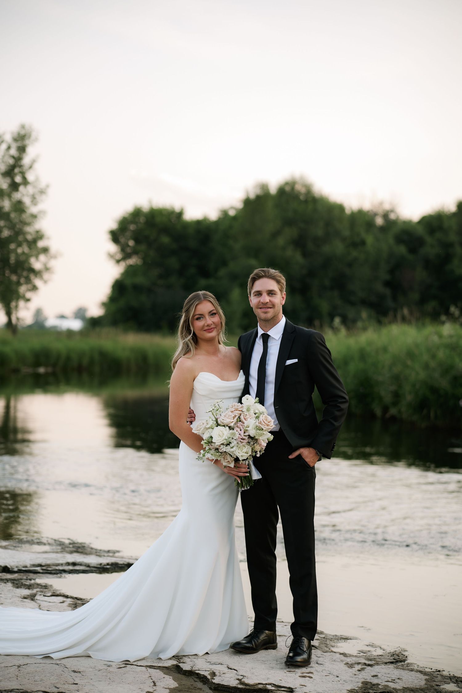 Bride and groom posing together at golden hour beside tranquil river surrounded by lush green trees.