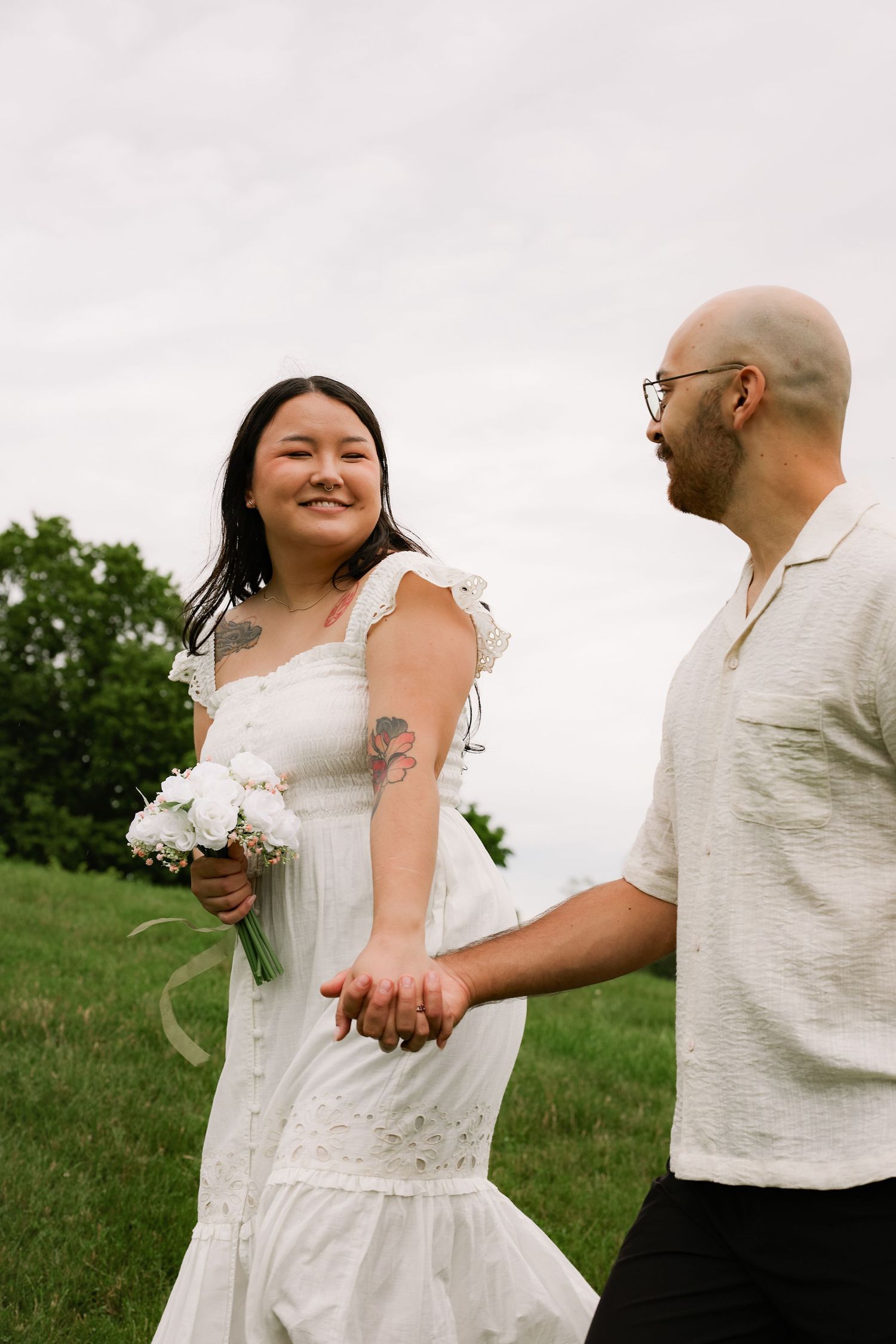 A smiling bride in a white dress holding flowers walks hand-in-hand with a bald groom in a field.