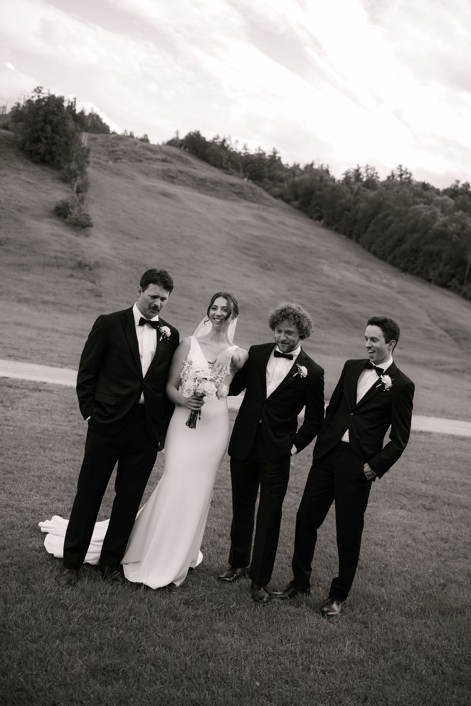 Black and white wedding photo of bride in white gown with three groomsmen in tuxedos on a hillside.