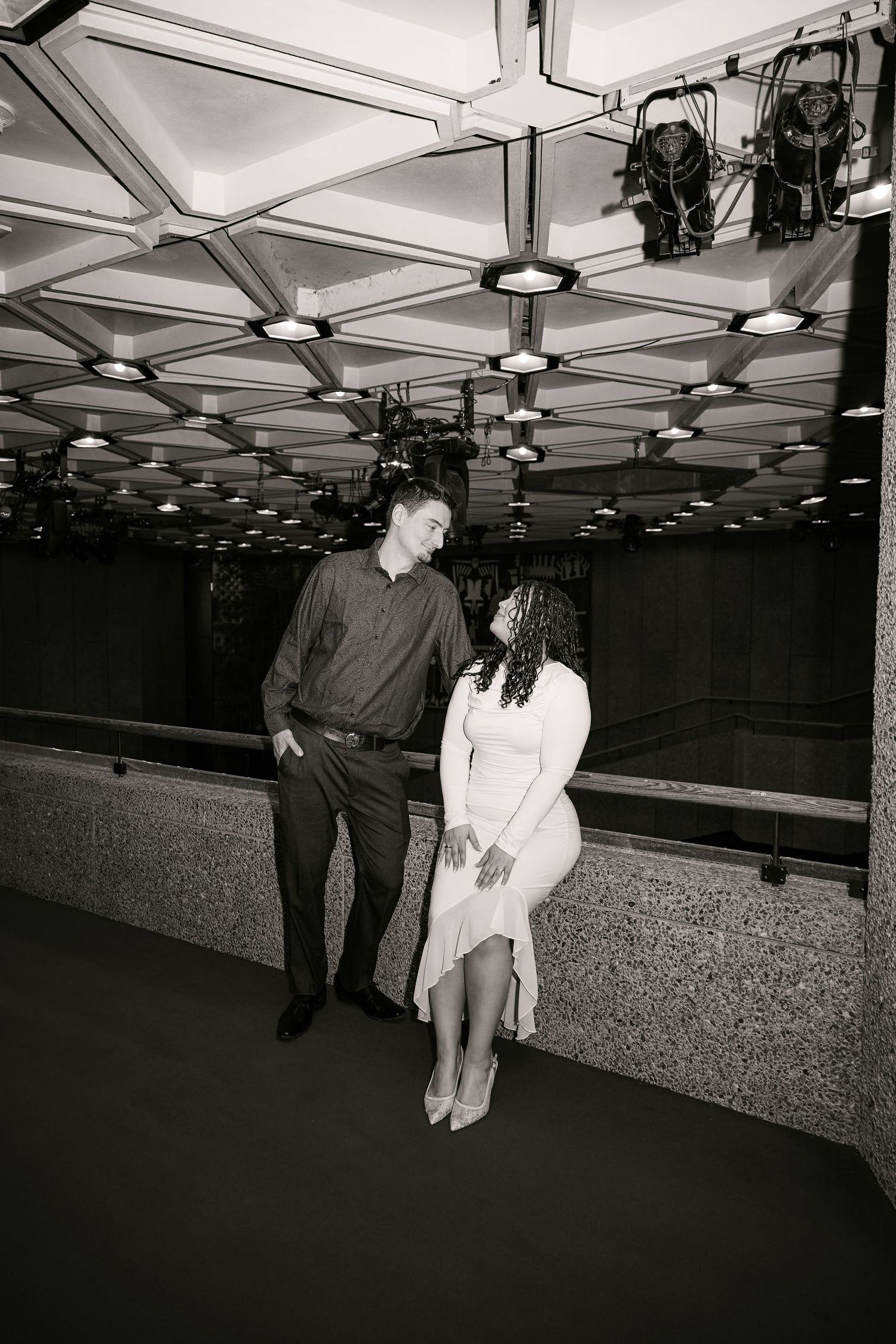 Black and white photo of a couple sharing a romantic moment under geometric ceiling lights in an elegant indoor space.