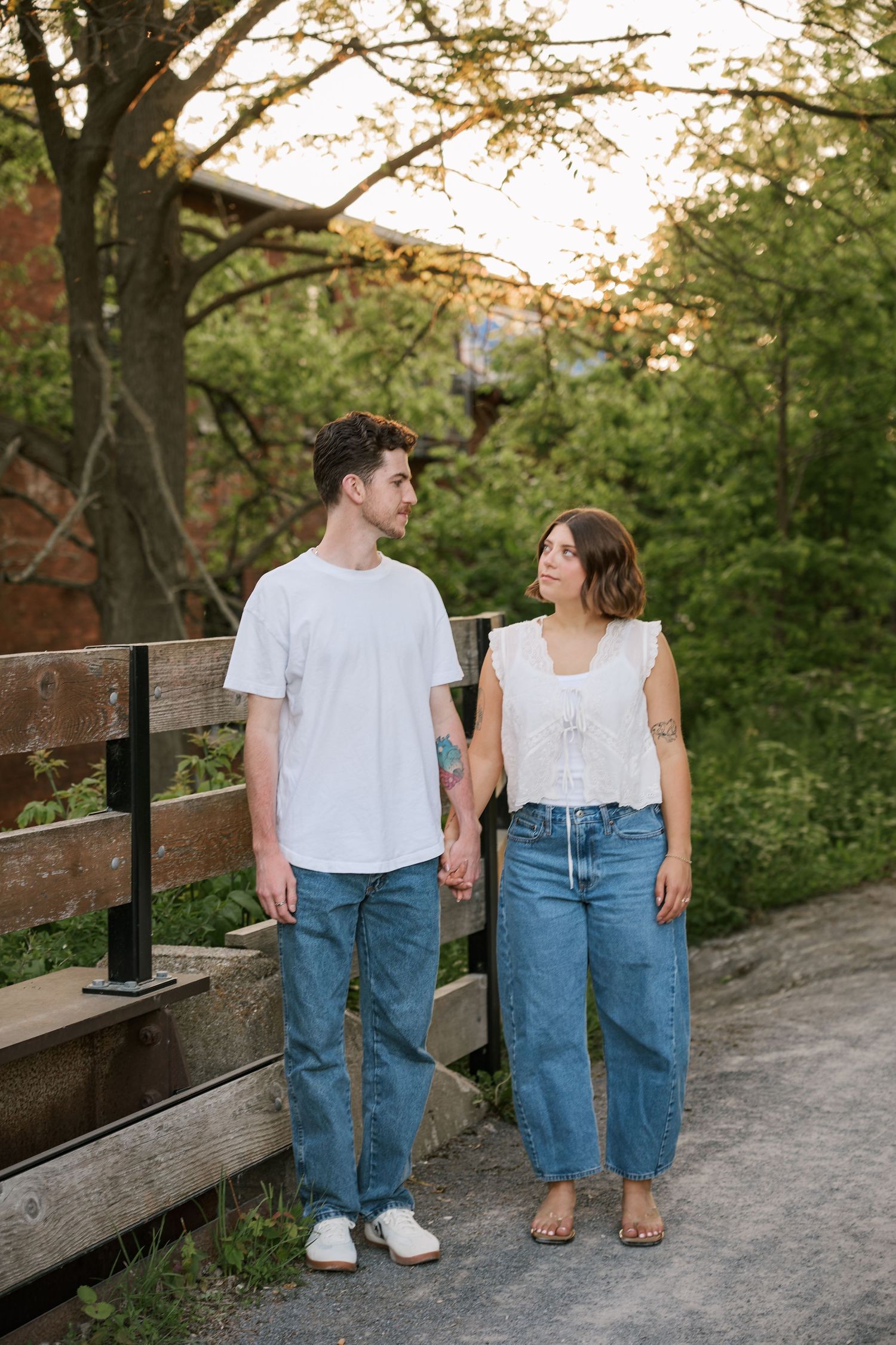 Couple holding hands and gazing at each other on a bridge path surrounded by lush green trees at golden hour.