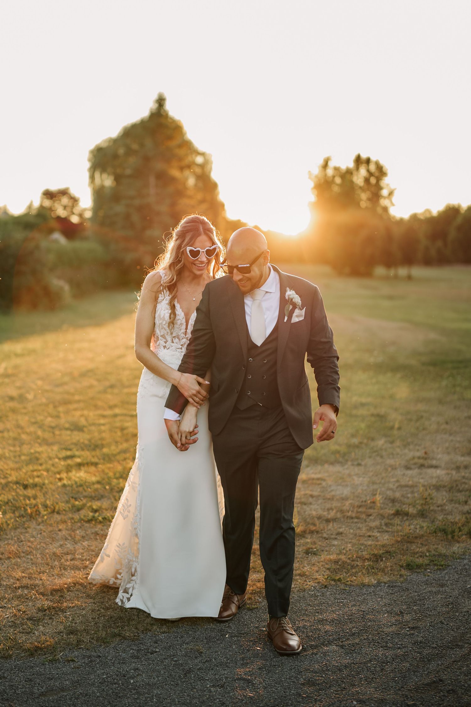 A bride and groom walk together at golden hour sunset, she wearing heart sunglasses and a white gown, he in a dark suit.