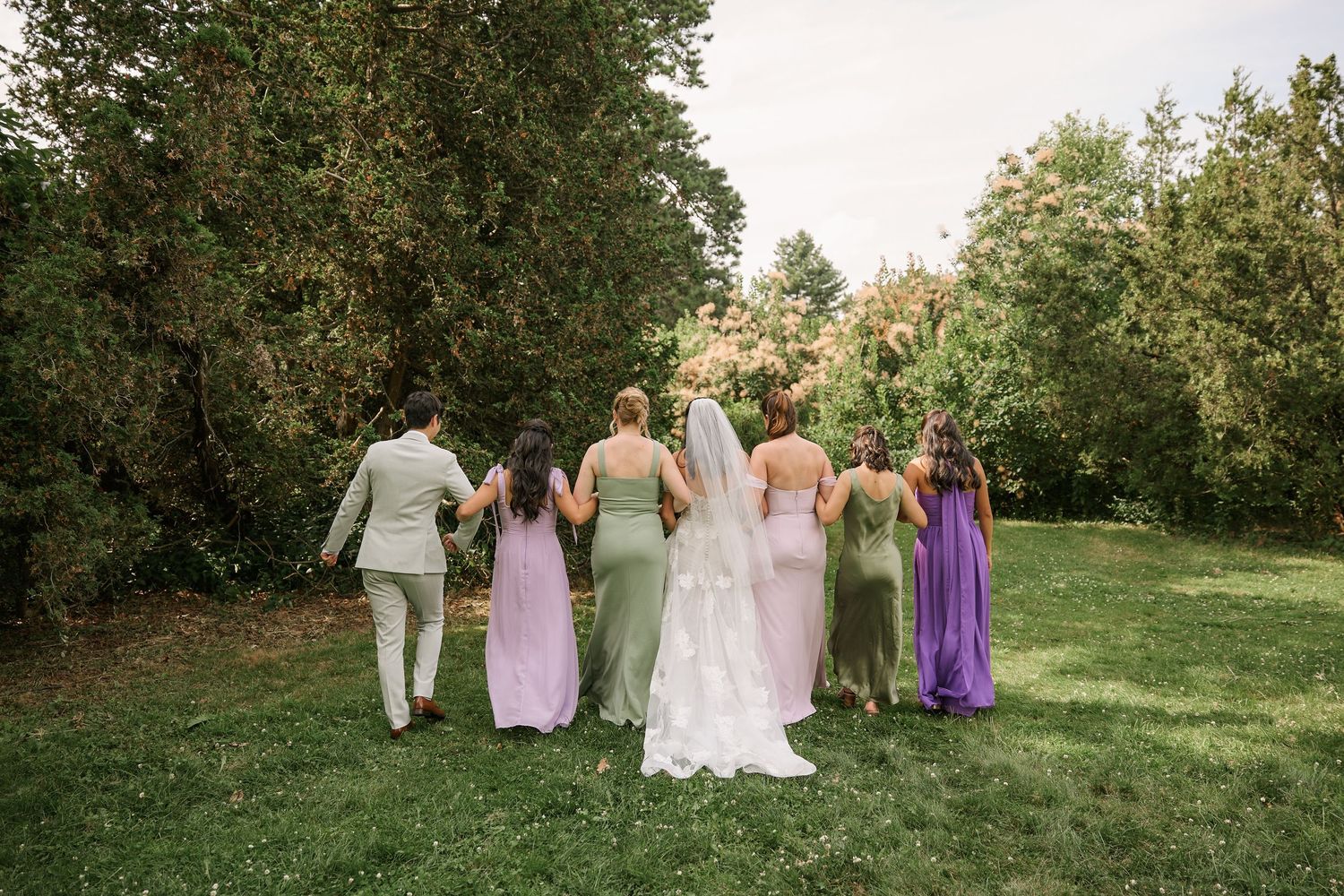 Wedding party walking together on grass path wearing pastel colored dresses.