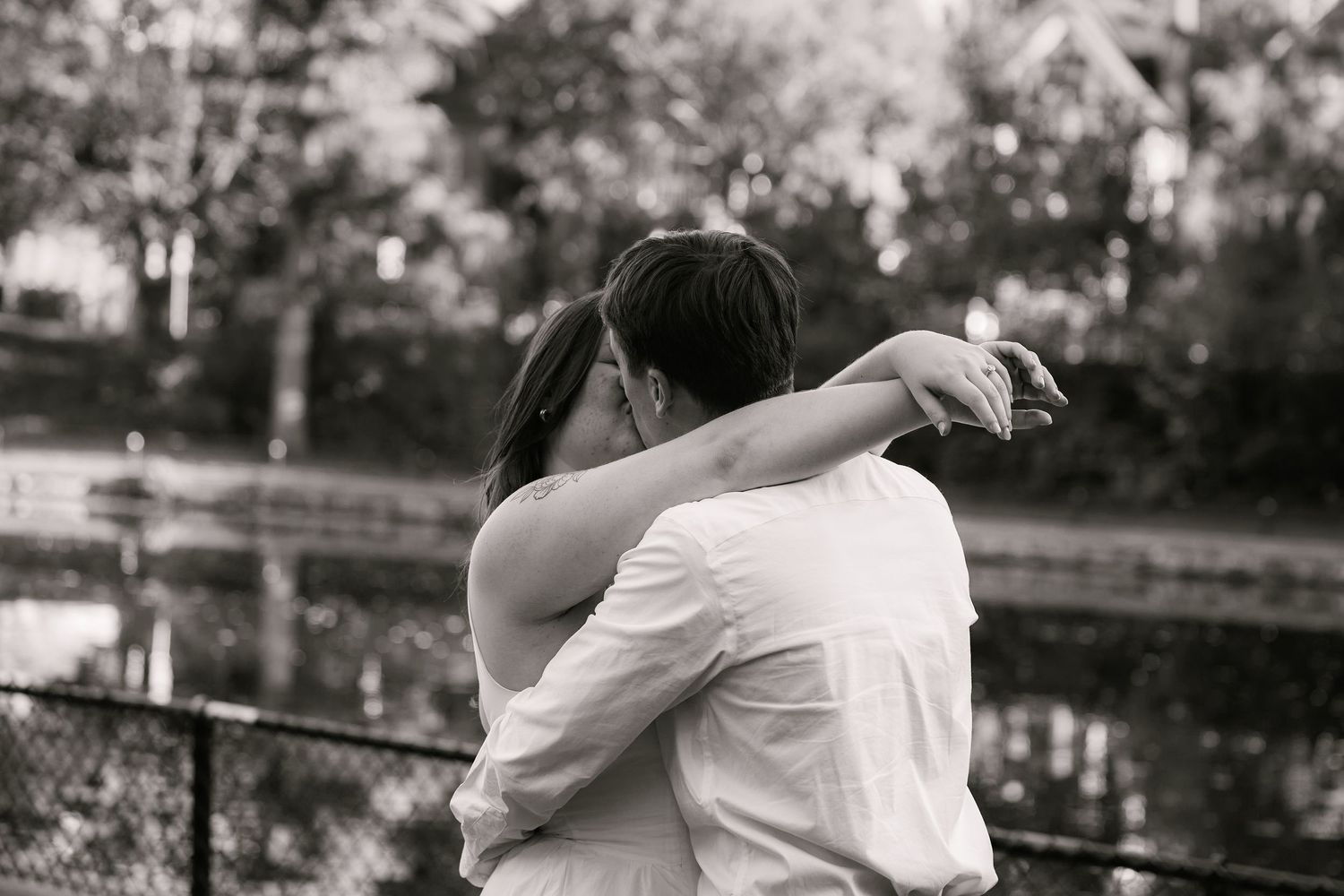 Black and white photo of a couple embracing and kissing near a fence with trees and water in the background.