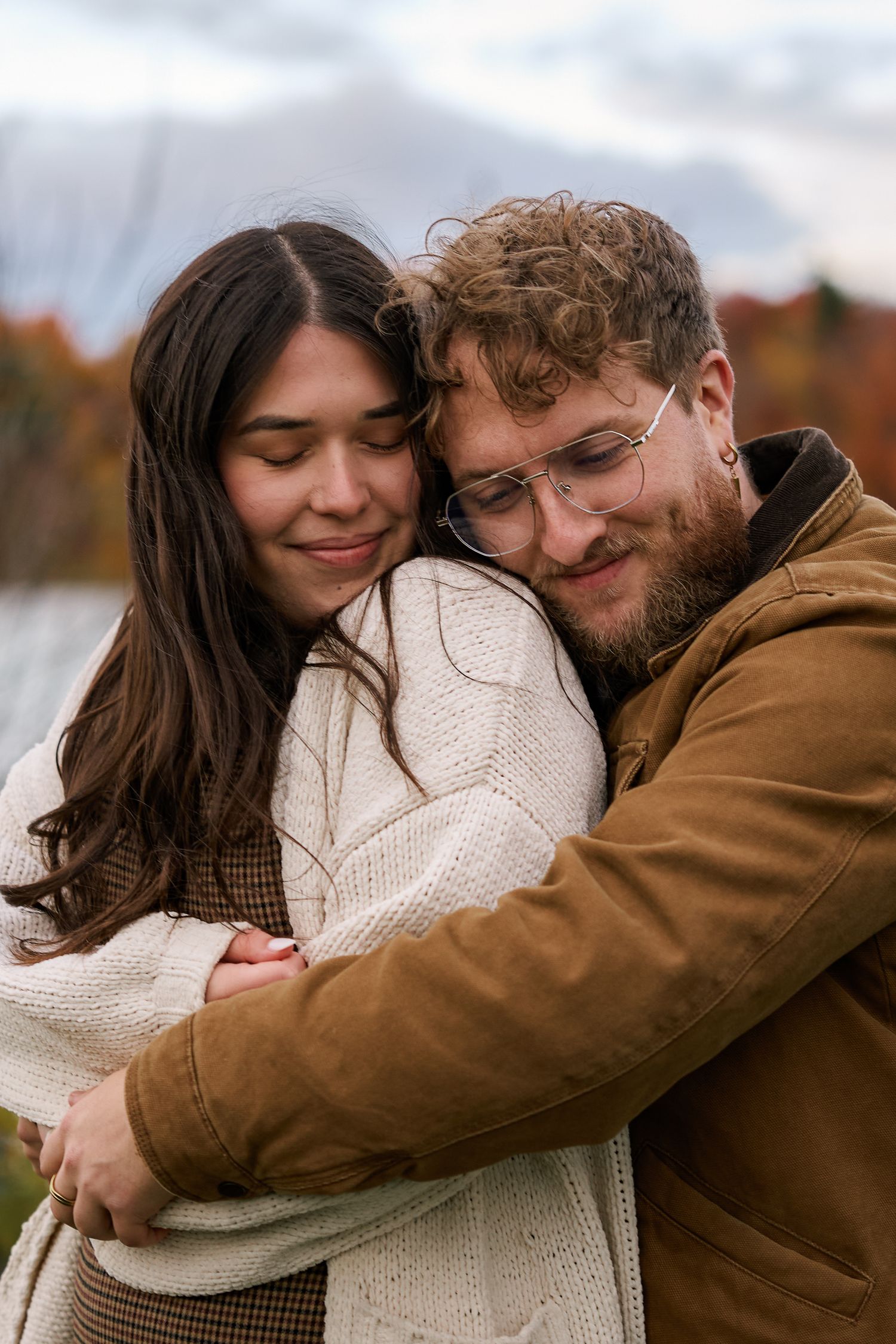 A couple embraces warmly outdoors in autumn, the woman in a white sweater leaning into a bearded man in a brown jacket.