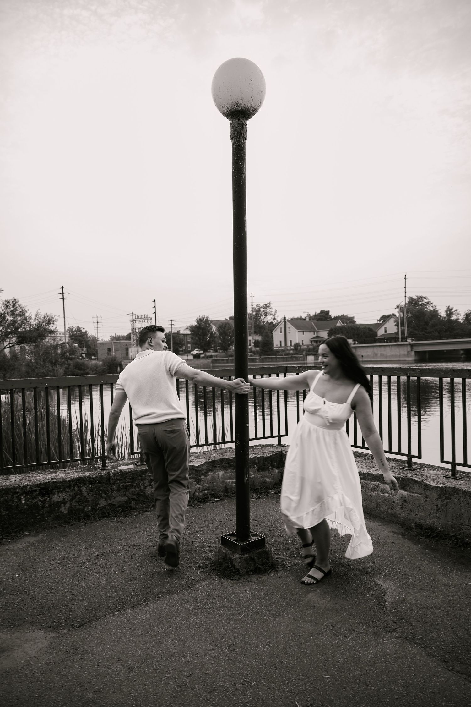 Black and white photo of a couple holding hands around a lamppost on a bridge near a river.