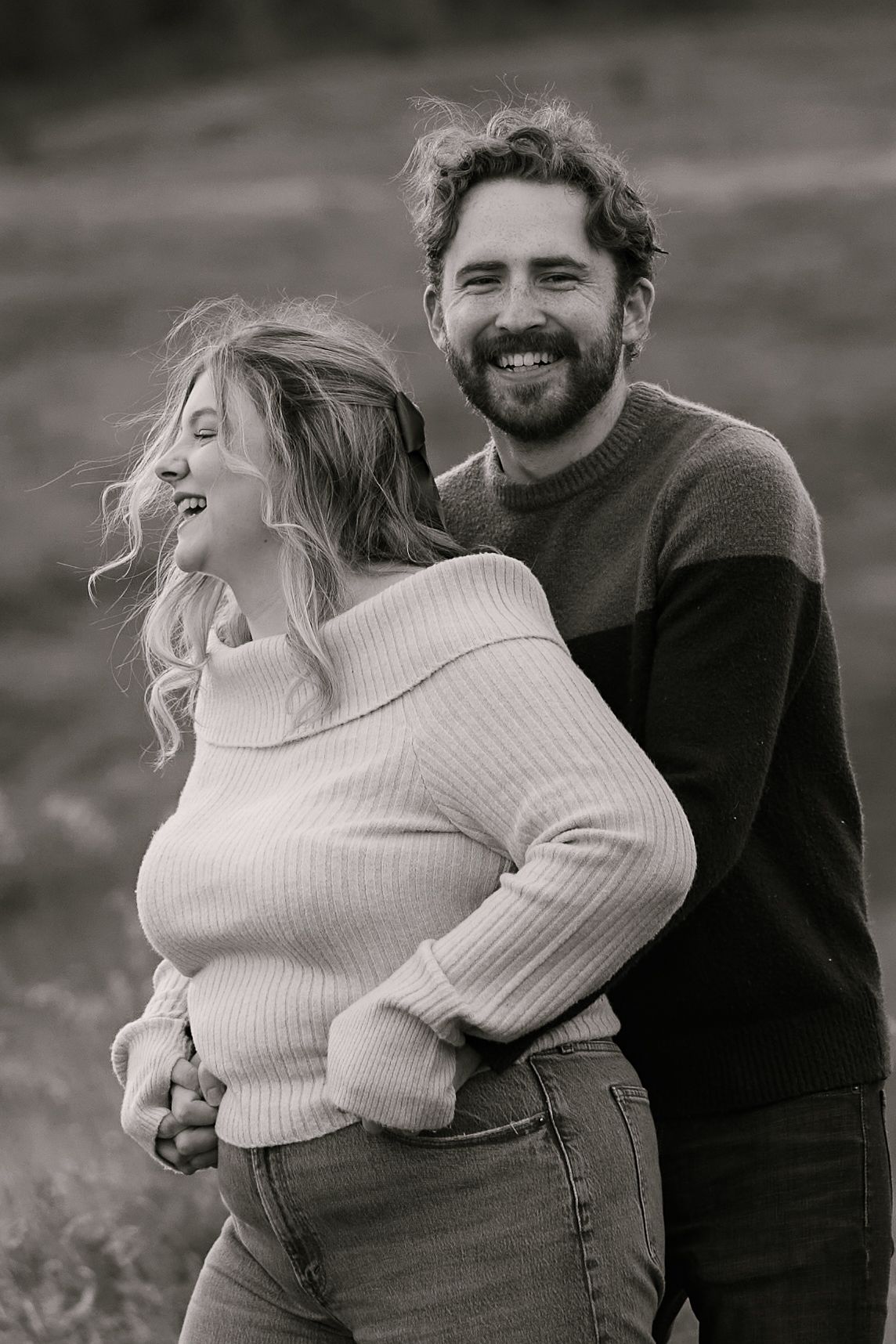Black and white photo of a smiling couple embracing outdoors, woman in off-shoulder sweater and jeans, man in colorblock sweater.