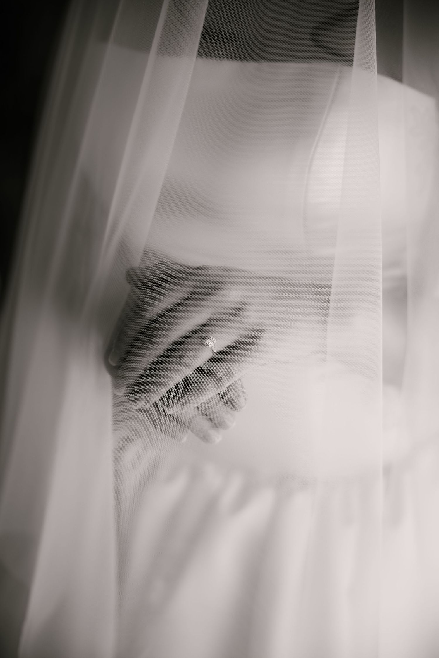 Close-up black and white image of hands with a wedding ring through delicate white fabric.