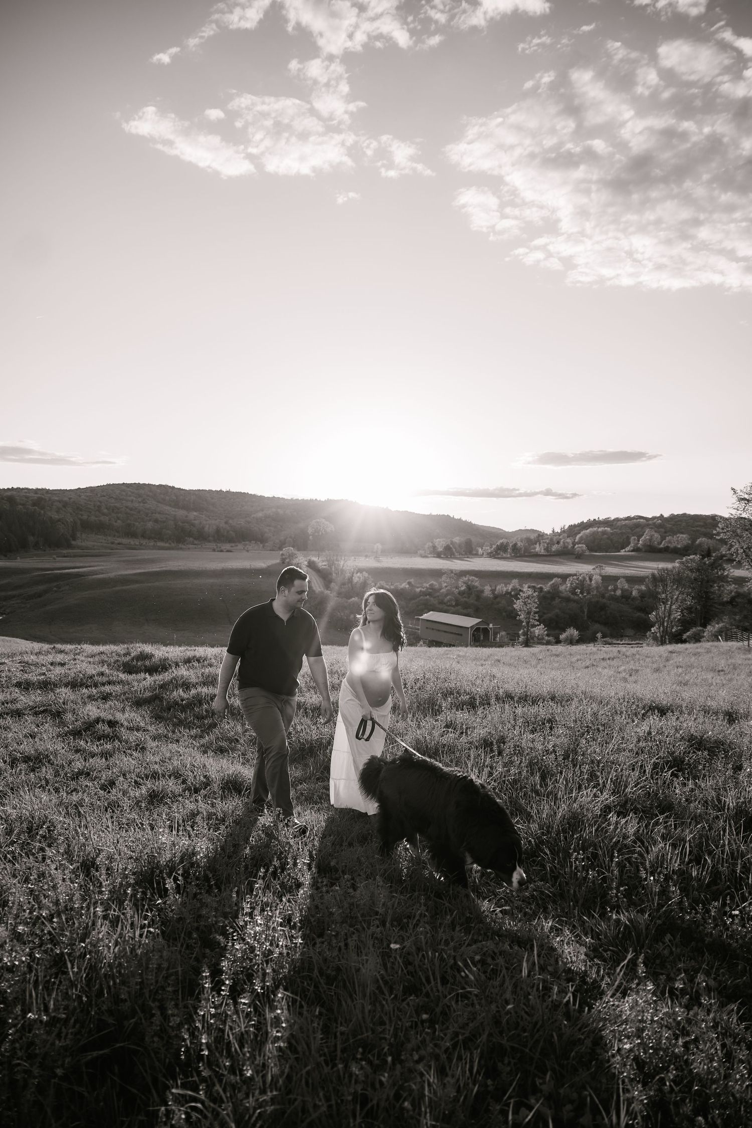 A couple walks with their dog through a meadow at sunset.
