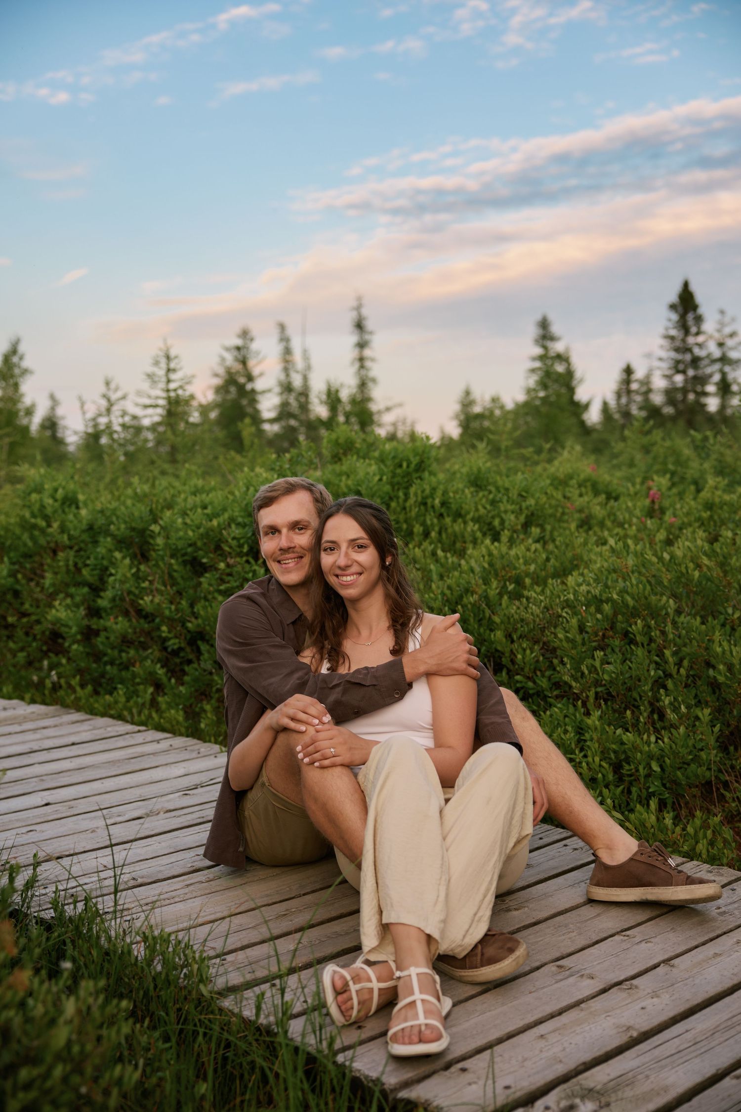 A couple lounges together on a wooden boardwalk surrounded by lush greenery at sunset.
