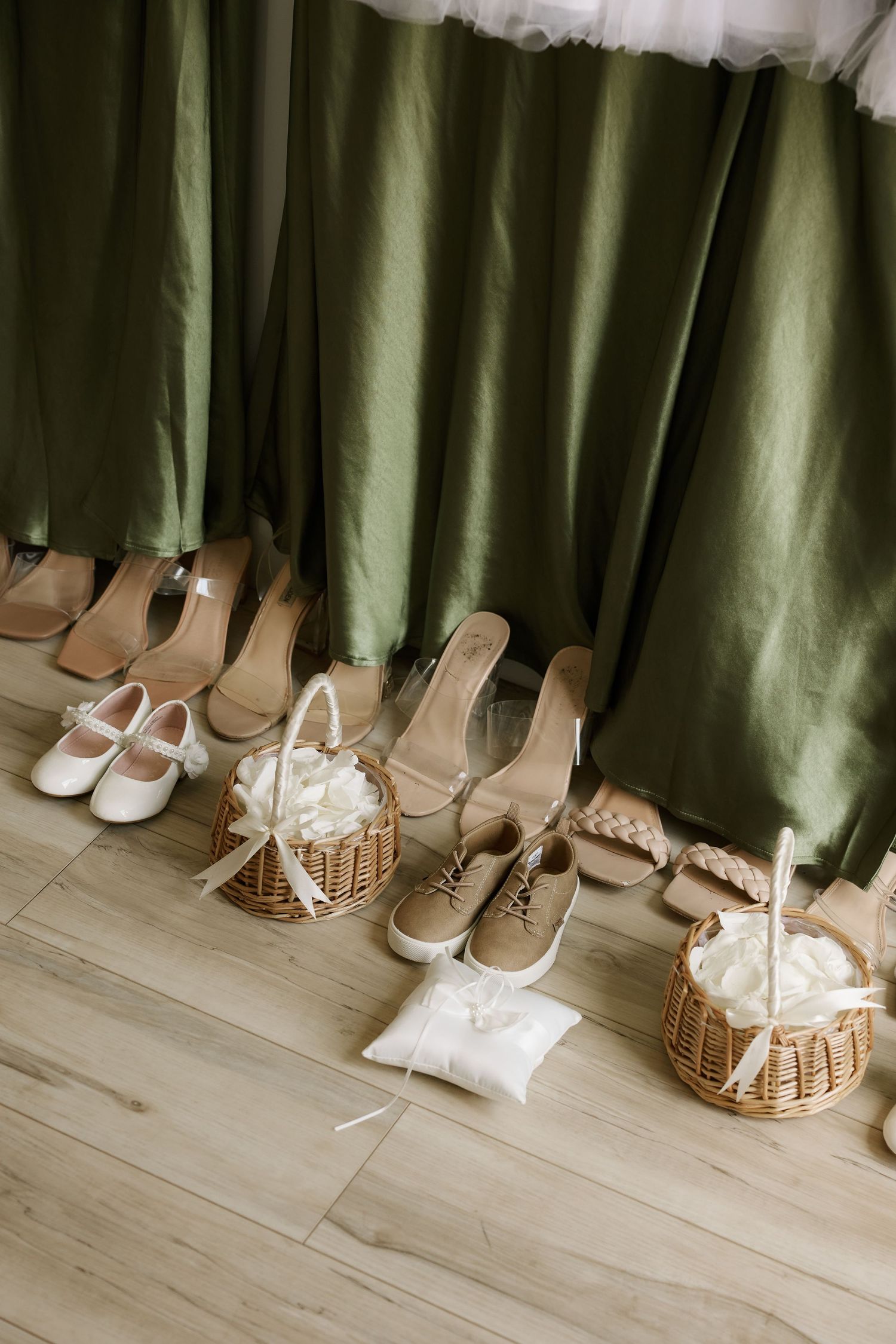 Wedding accessories arranged on wood floor including bridal shoes, flower girl baskets, and ring pillow beside green bridesmaid gowns.