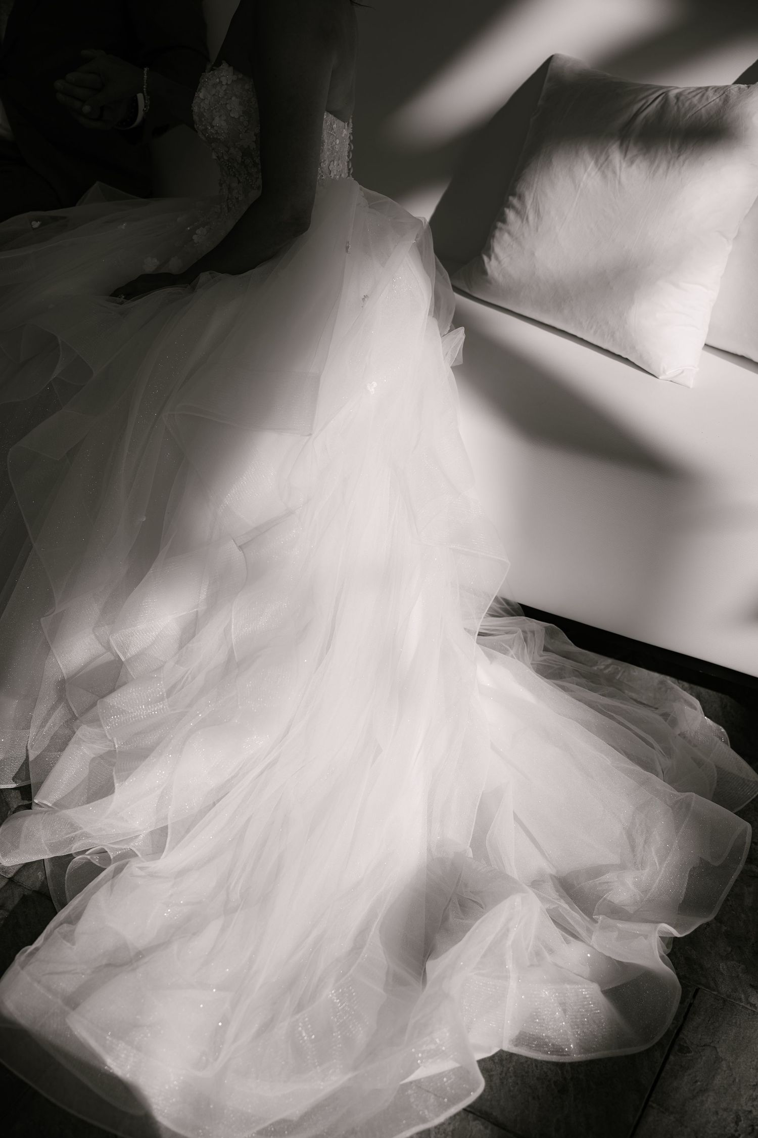 Black and white photo of a bride seated on a sofa, her layered tulle wedding gown flowing across the floor beside a soft pillow.