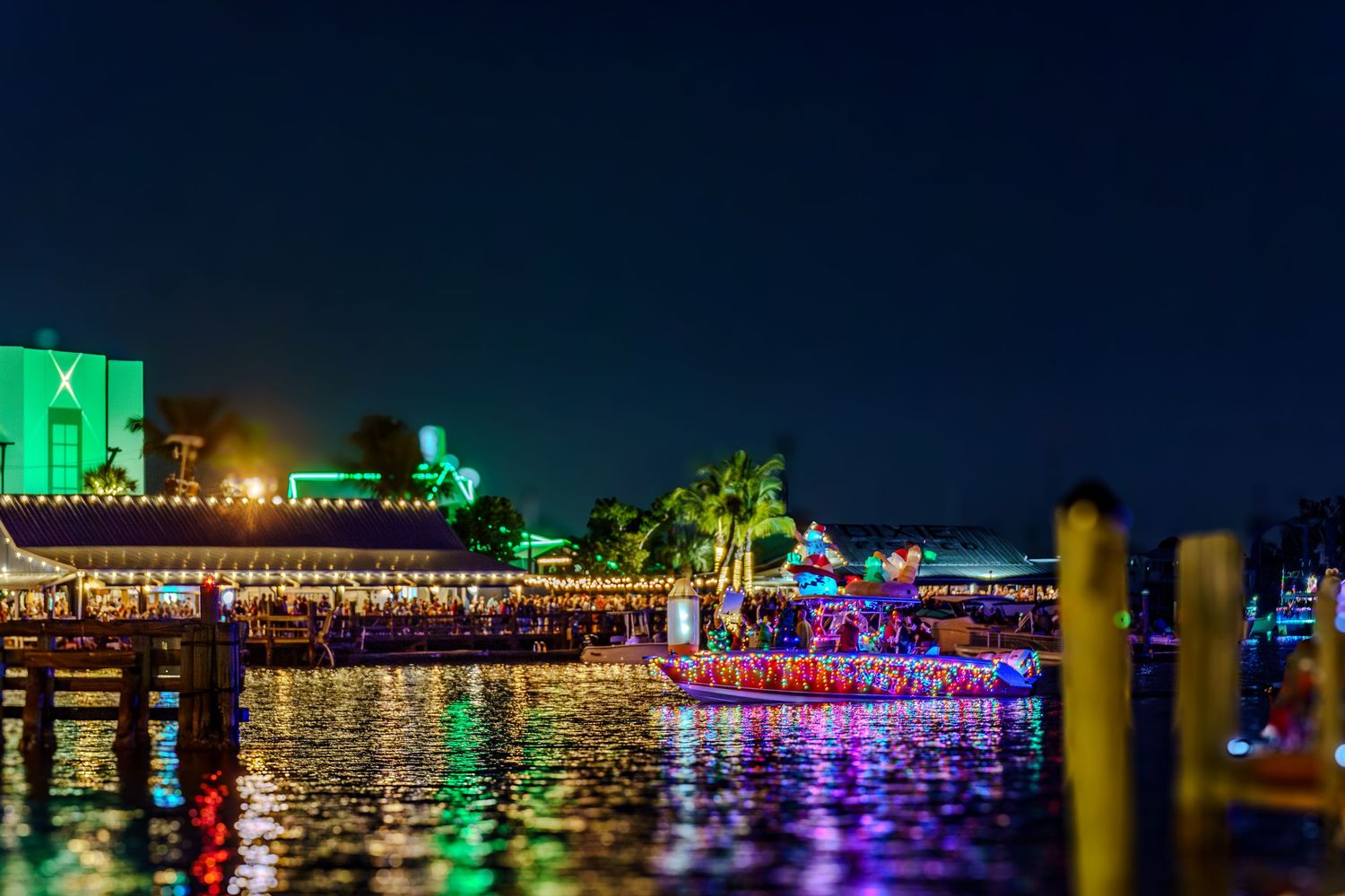 Colorful LED lights illuminate a waterfront boardwalk and buildings reflecting on calm water at night.