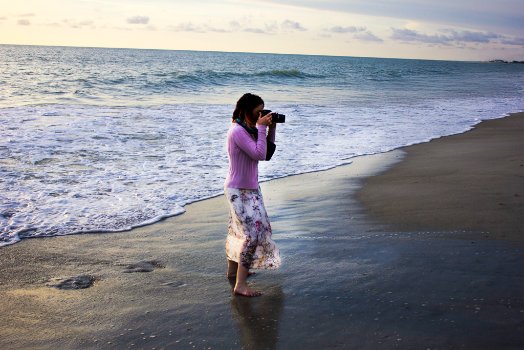Photographer in pink shirt taking pictures along a sandy beach at dusk.