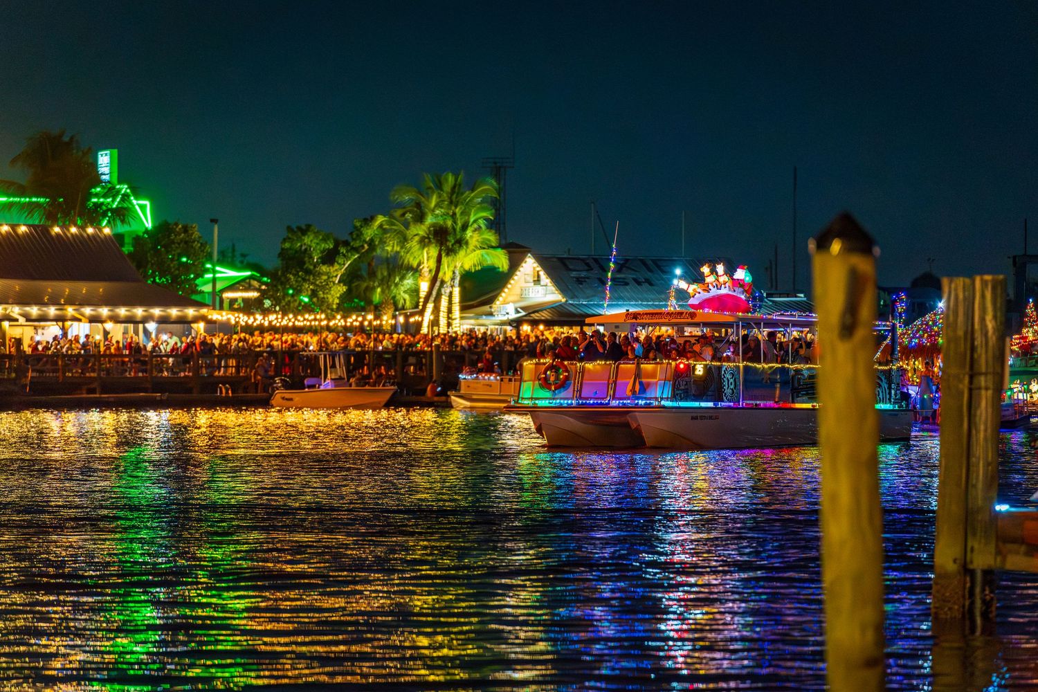 Colorful lights illuminate boats and buildings along a waterfront at night creating a vibrant reflection on the water.