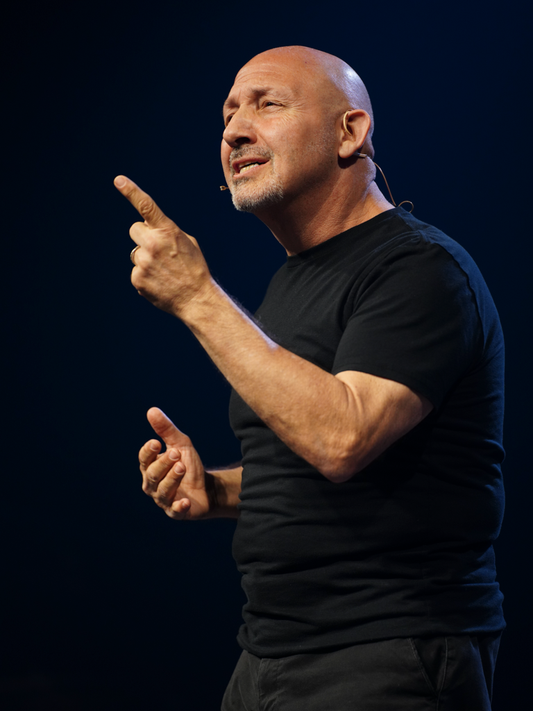 Speaker in black t-shirt gesturing animatedly against dark background.