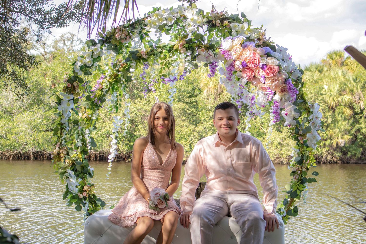 A couple sits under a floral arch by a waterway during an outdoor photoshoot.
