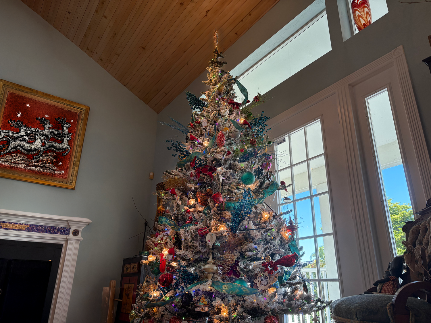 Festive Christmas tree with colorful ornaments and lights in a cozy room with wood ceiling and windows.