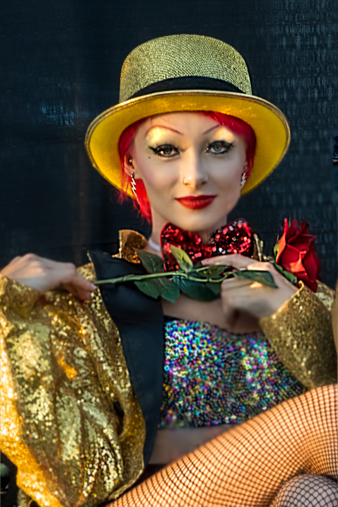 Glamorous cabaret performer in gold sequins and yellow hat poses dramatically against dark background.