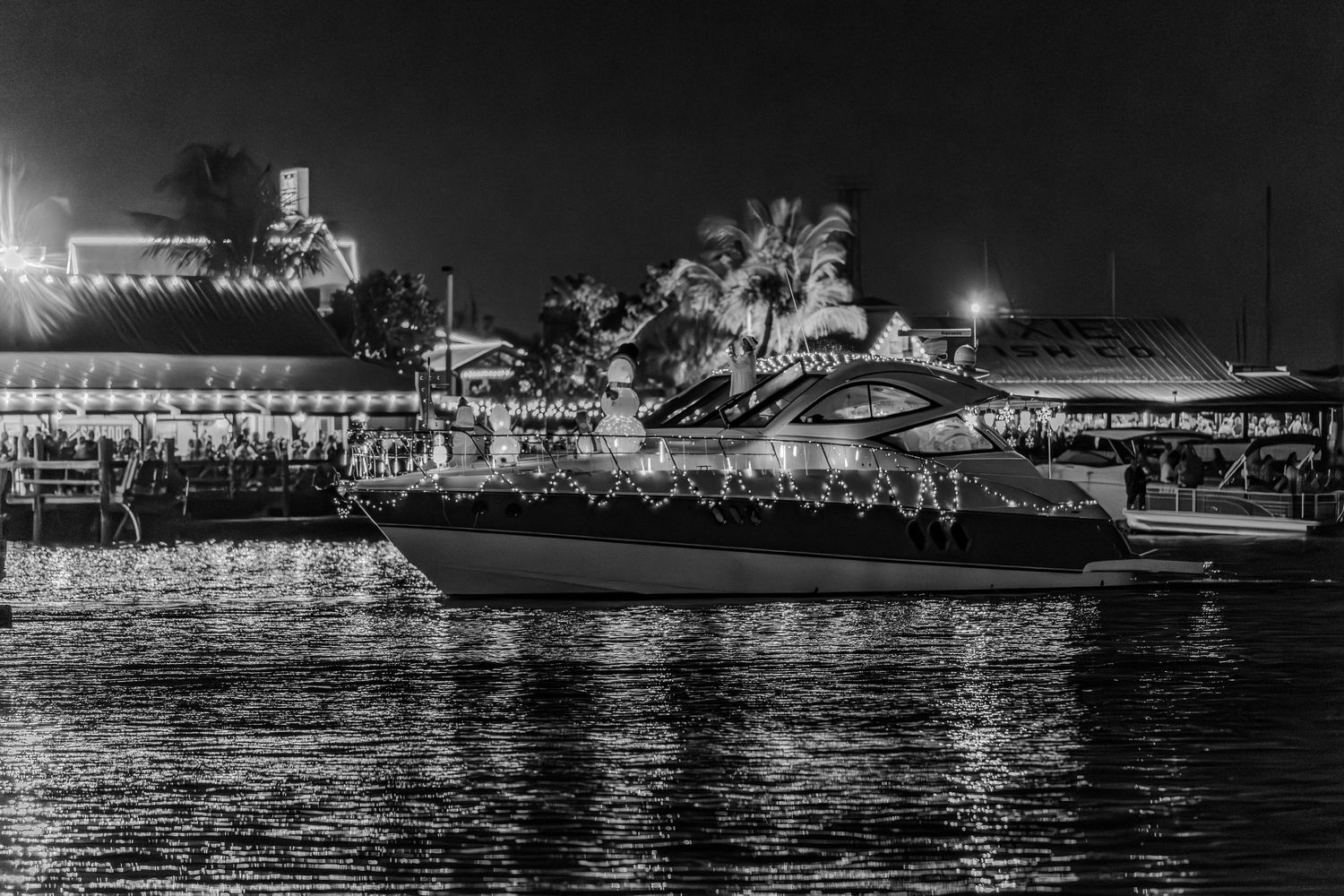 Illuminated boats and waterfront buildings create a dramatic black and white nighttime reflection on calm waters.