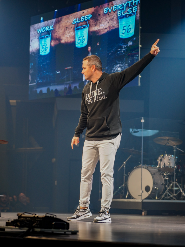 Speaker on stage in black jacket and light pants gestures energetically during presentation.