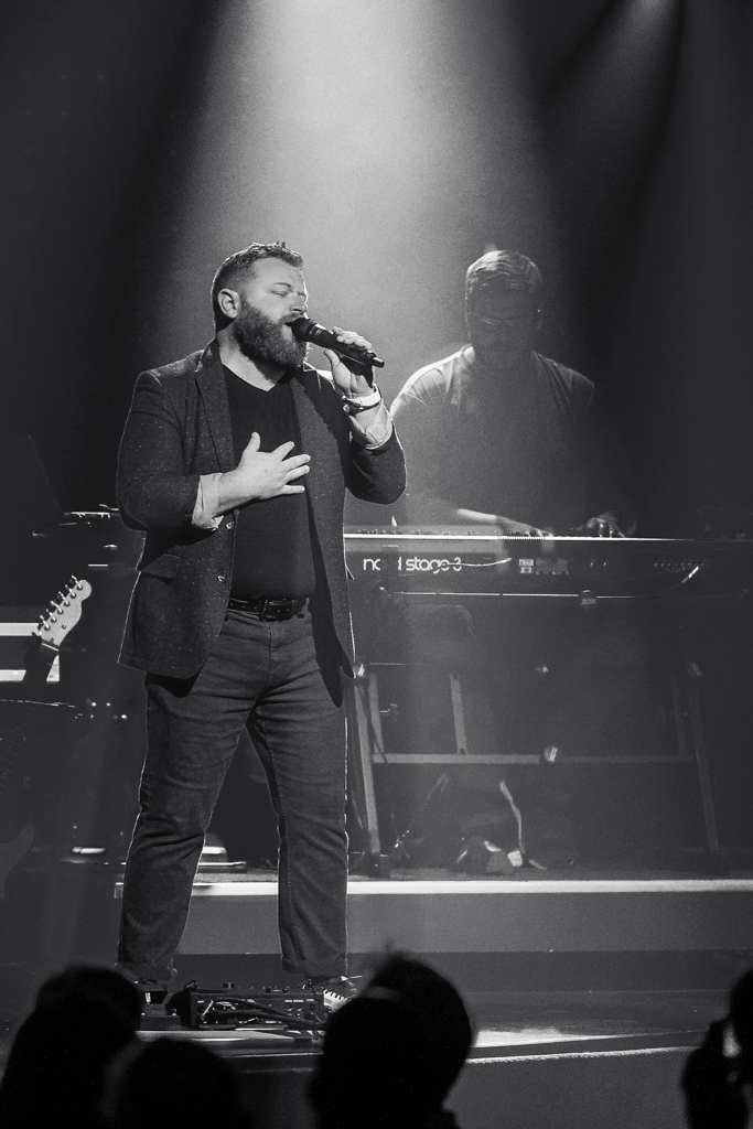 Concert performer in dark clothing sings passionately on dimly lit stage.
