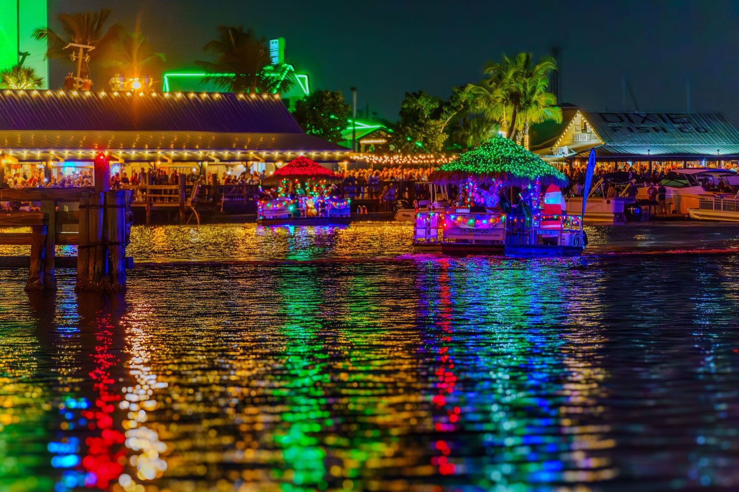 Colorful holiday lights reflect on calm water at night with illuminated docks and buildings along the shoreline.