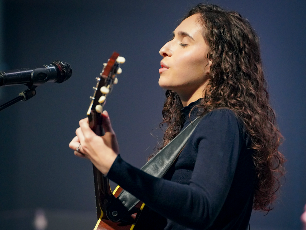 Musician in black attire plays guitar against dark blue backdrop on stage.