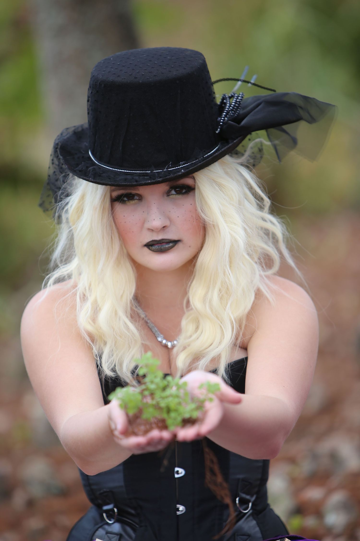 Gothic-styled figure in black attire and top hat poses with plants outdoors.
