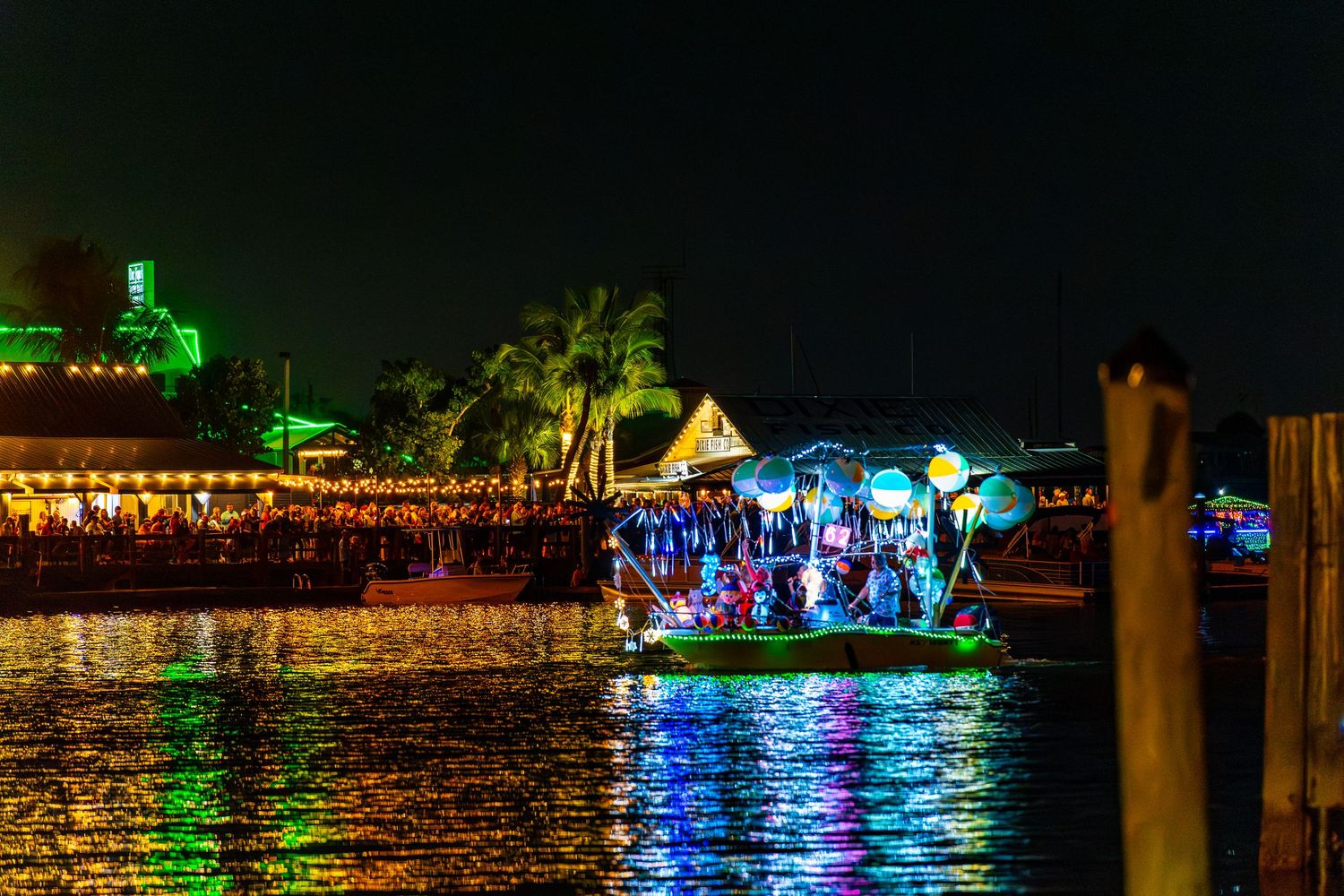 Colorful holiday lights reflect off calm waterfront with illuminated buildings and boats creating a festive nighttime scene.