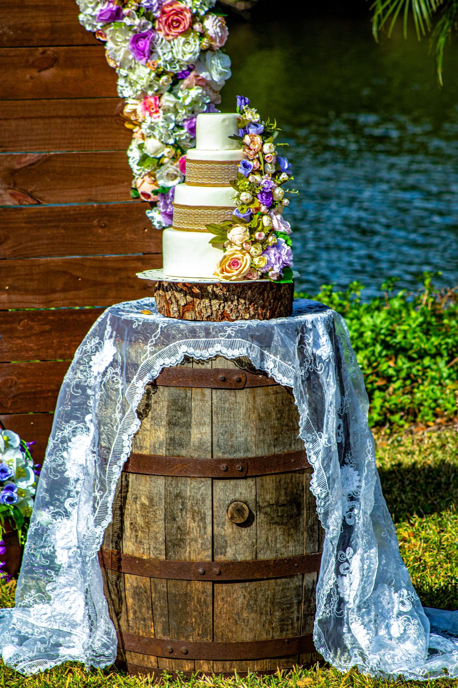 Wedding cake display on rustic wooden barrel with floral decorations and lace accents.