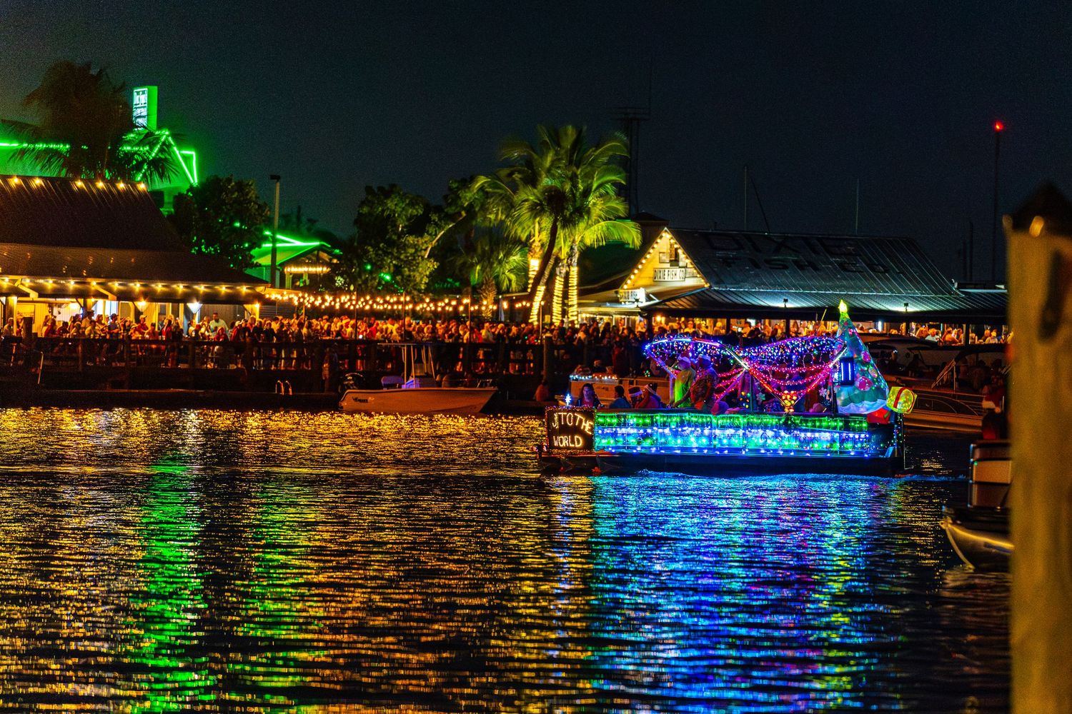 Colorful nighttime waterfront scene with illuminated boats and buildings reflecting on dark water during a festival.
