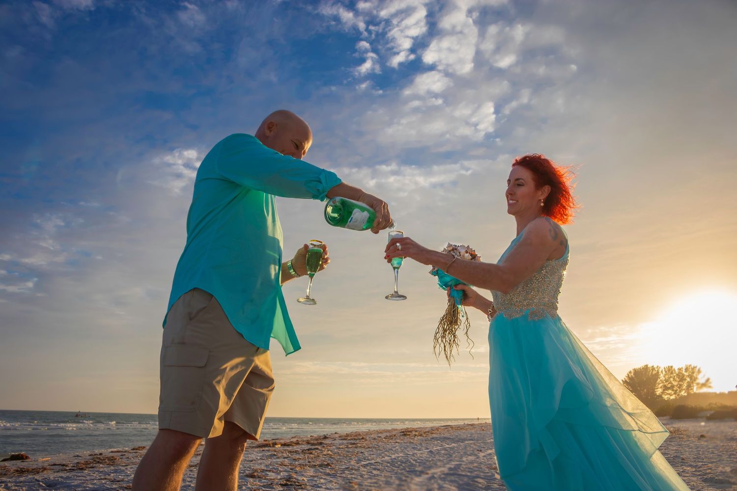 A couple in turquoise outfits dances playfully on the beach at golden hour.