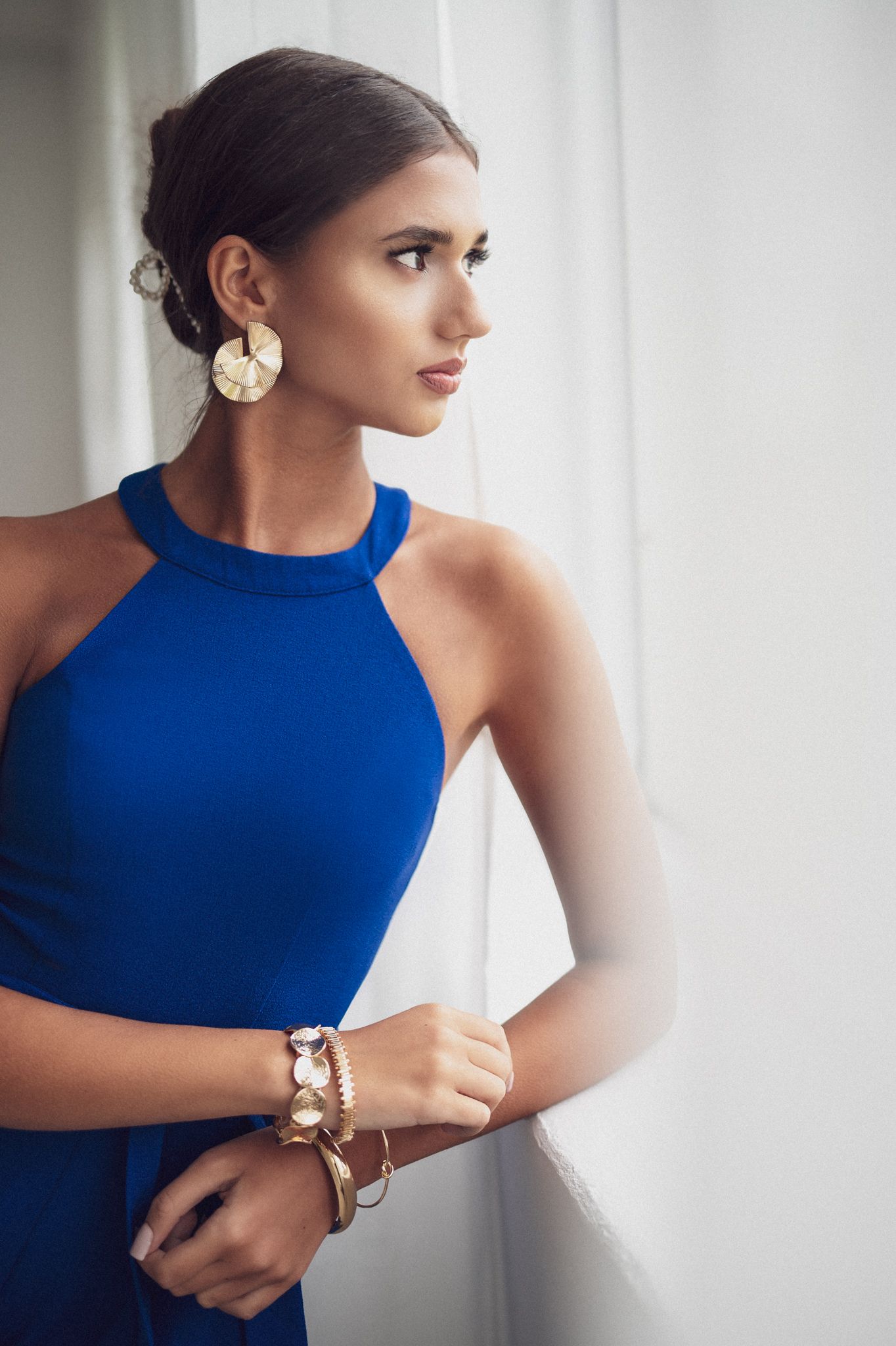 Elegant portrait with model in royal blue high-neck top wearing gold accessories against white backdrop.