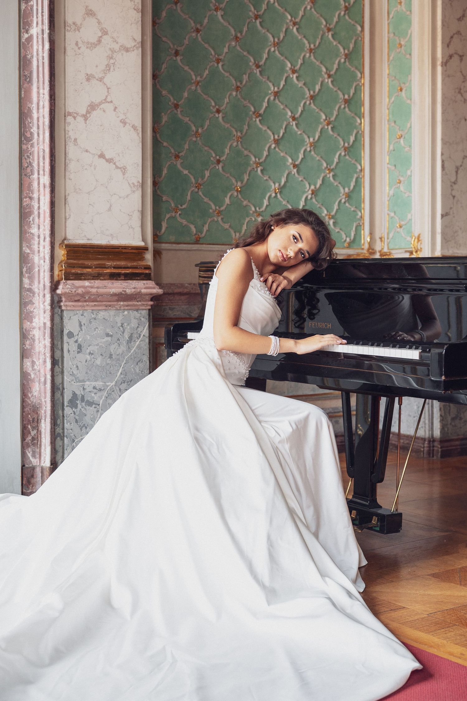 Elegant bride in white gown resting on a grand piano in an ornate room with green walls.