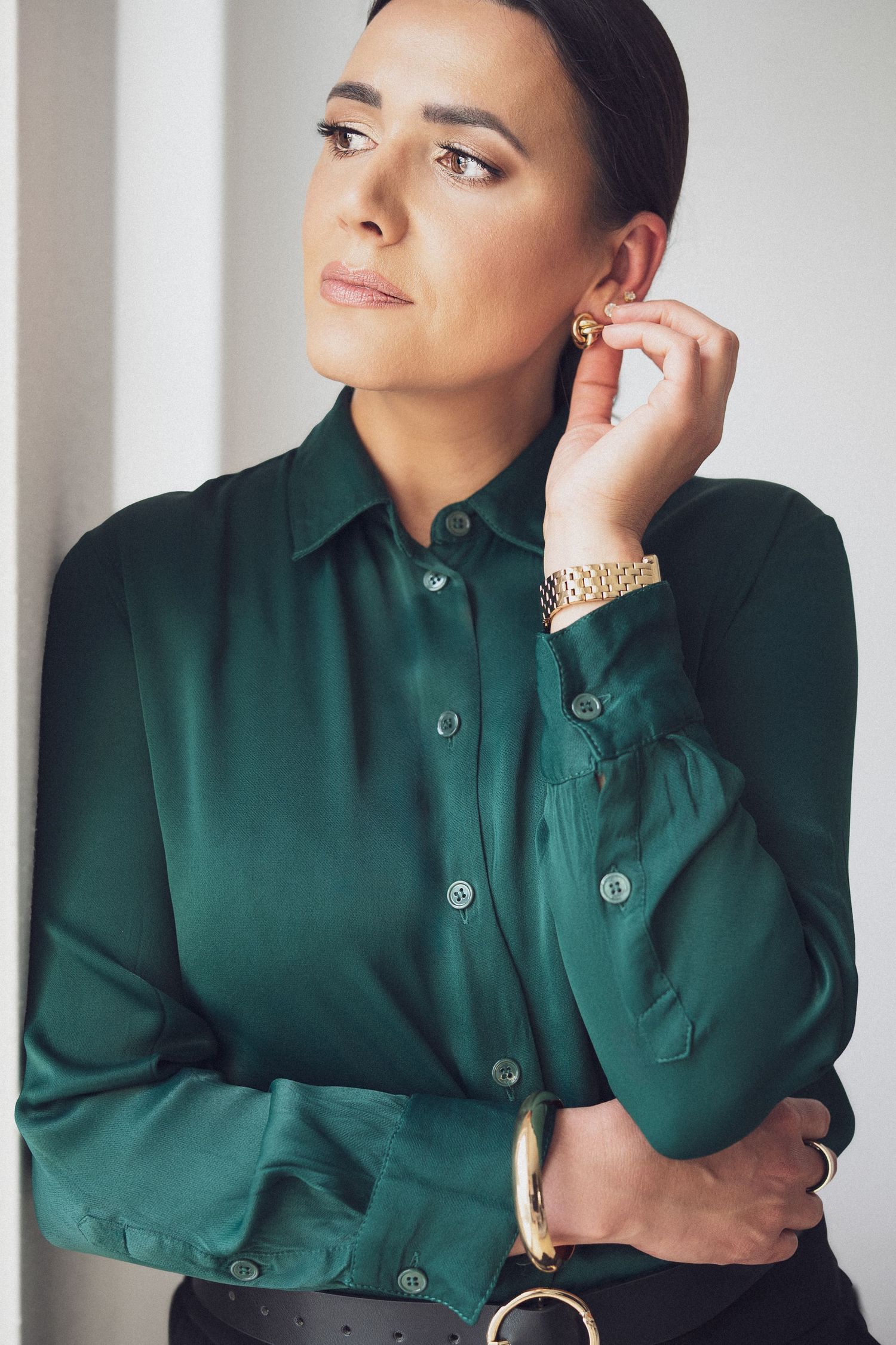 Woman in green silk blouse with gold earrings and watch, posing thoughtfully against a white wall.